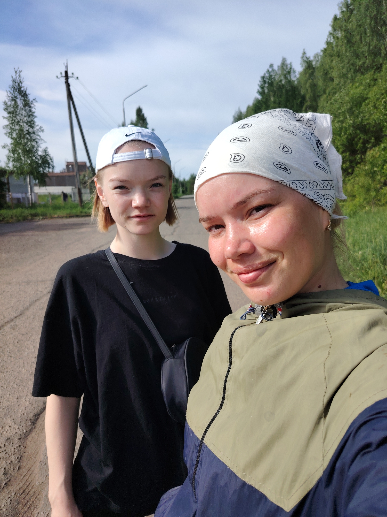 Two young women pose for a selfie on an asphalt road in Марковский, Russia. The woman in the foreground wears a white bandana, a green and navy blue jacket, and small gold earrings. Her skin appears slightly shiny, possibly due to perspiration. Behind her and to the left, the other woman wears a white baseball cap with a black logo, a black t-shirt, and a black shoulder bag. They stand on a road that shows signs of wear and tear, with cracks and patches. The background includes a utility pole, streetlights, trees, and building. The sky is partly cloudy.