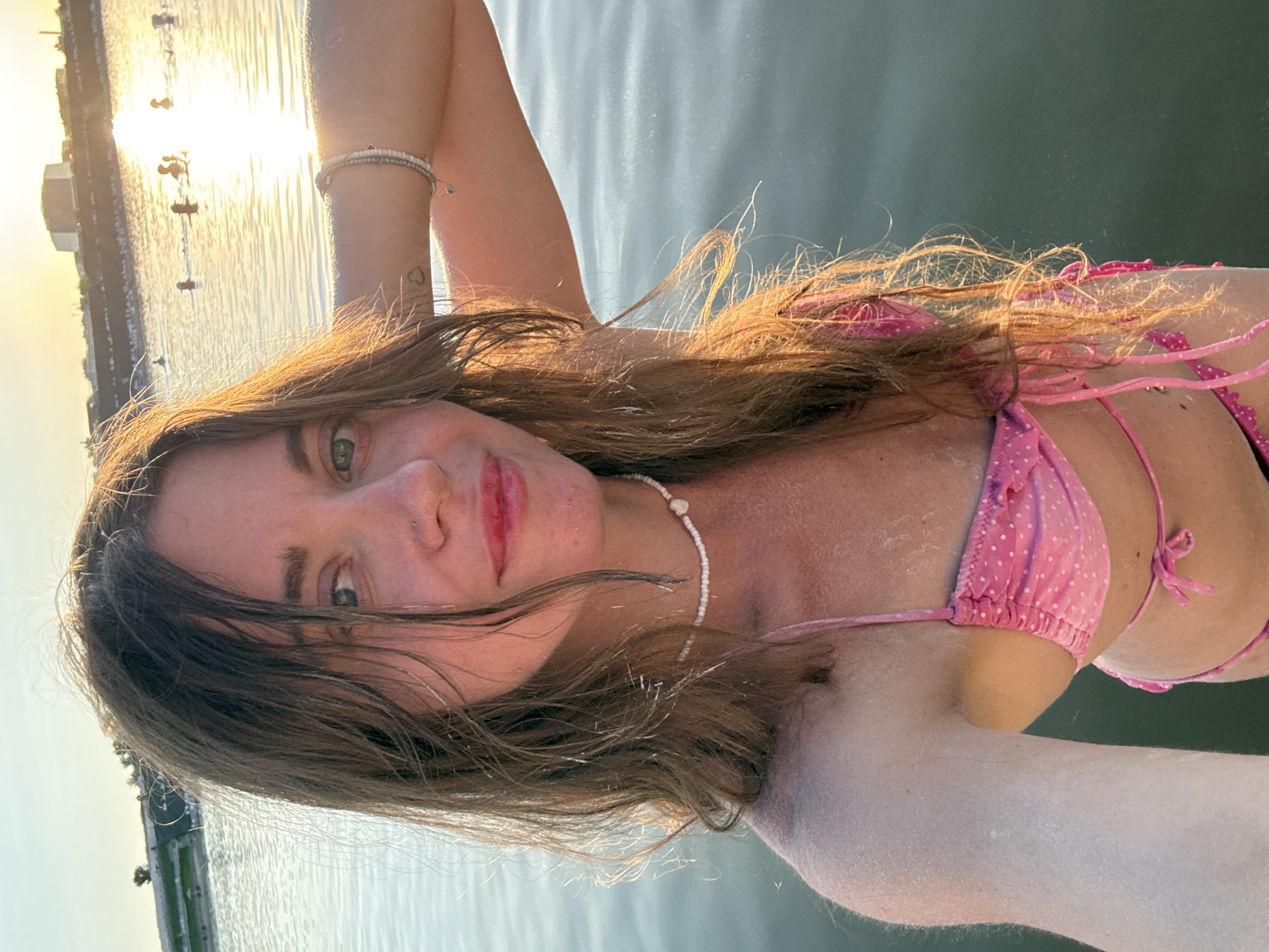 In Sol-Iletsk, Russia, a woman with long brown hair is captured in a selfie at eye level, set against the backdrop of a body of water. She is wearing a pink polka-dot bikini and a white beaded necklace. Her right arm is raised behind her head, adorned with bracelets. The water, illuminated by the warm glow of the setting sun, reflects the golden hues of the sky. In the distance, several people are visible in the water, and a building complex is situated on the far shore. The scene conveys a serene and tranquil atmosphere.