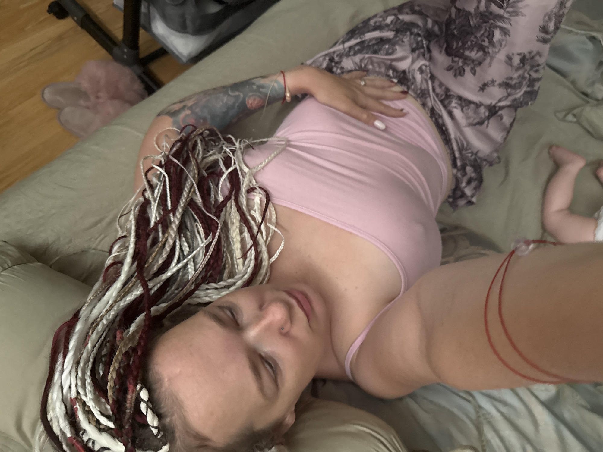A woman with blonde and red braided hair is lying on a bed with a baby's legs visible near the bottom of the frame. She is wearing a pink tank top and patterned pajama pants. The woman has tattoos on her arm and is wearing jewelry on her wrist and finger. The environment appears to be a bedroom with wooden flooring visible in the background, along with pink fuzzy slippers and a vacuum cleaner. There is no indication of the location being Moscow, Russia.