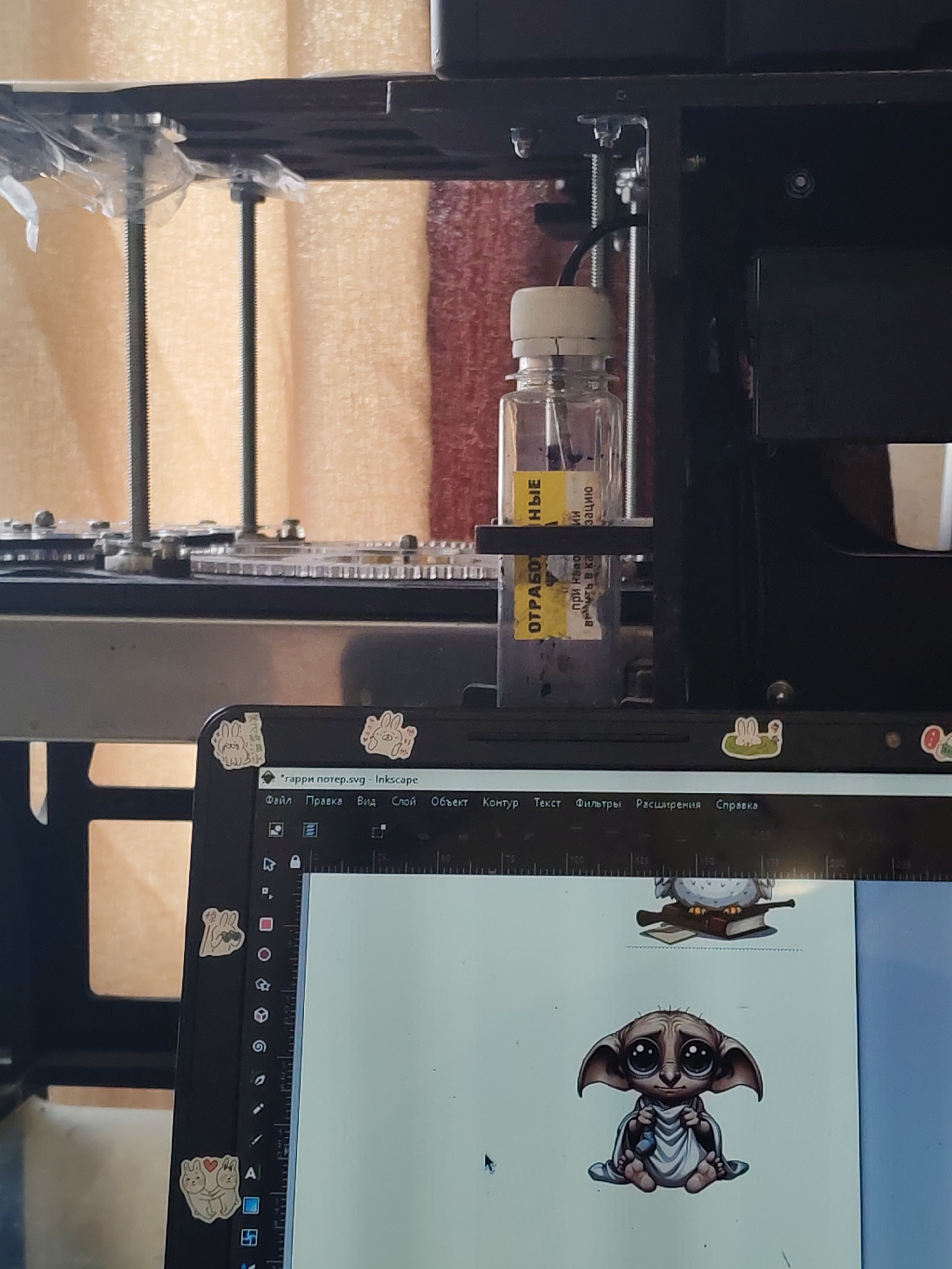 The scene presents a close-up view of a workspace, likely indoors, dominated by technical equipment and a laptop. In the background, a machine with metallic components and visible vertical threaded rods is partially visible. A small, clear plastic bottle containing a dark liquid is attached to the machine, with a yellow label displaying text in Russian. The label includes the word "ОТРАБОТ" followed by more text. The machine is positioned against a background consisting of light and dark fabric, possibly curtains.
In the foreground, a laptop screen displays an Inkscape interface. The file name "гарри потер.svg" is visible in the Inkscape title bar. The screen shows a graphic design featuring a depiction of Dobby from the Harry Potter series and another image of an owl sitting on books. Several stickers are adhered to the laptop, including bunny and peace sign designs. The environment is dimly lit, with a mixture of natural and artificial light.