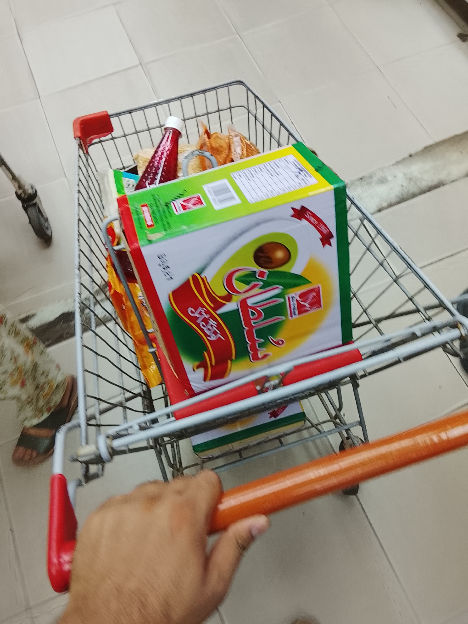 A hand holds the handle of a shopping cart, which is partially filled with groceries, including a large box with Arabic writing, a bottle of red sauce, and other items. The location appears to be a store aisle, based on the tiled floor. No specific indicators within the image confirm the location within Gulgasht Colony, Pakistan.