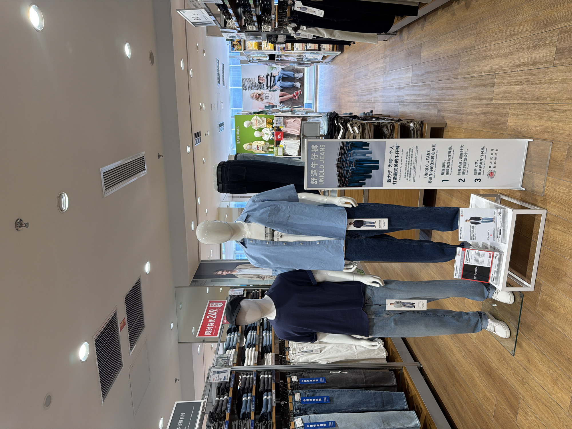 The image depicts the interior of a clothing store, likely a Uniqlo, located in Qinhuai District, China. The scene is well-lit with recessed ceiling lights and spotlights illuminating the retail space. Wooden flooring runs throughout the store, and displays of jeans and casual wear are prominent.
In the foreground, two mannequins are dressed in casual outfits. The mannequin on the left wears a dark blue t-shirt and light blue jeans, topped with a black baseball cap. The mannequin on the right is clad in a light blue button-down shirt and dark blue jeans. Both mannequins are standing on clear acrylic stands.
To the left of the mannequins, shelves are stocked with folded jeans, arranged by size and style. A prominent red and white sign advertising a limited-time offer for 249 yuan is visible. To the right of the mannequins, a white display table holds promotional materials, including a sign with the Uniqlo Jeans logo and product information in Chinese and English. Further to the right, racks of clothing, including a selection of dark colored garments, are displayed.
In the background, a large advertising poster features images of children and young adults modeling Uniqlo clothing. Beyond this, the store continues with more merchandise displays and walkways. The overall impression is that of a clean, organized, and modern retail environment.