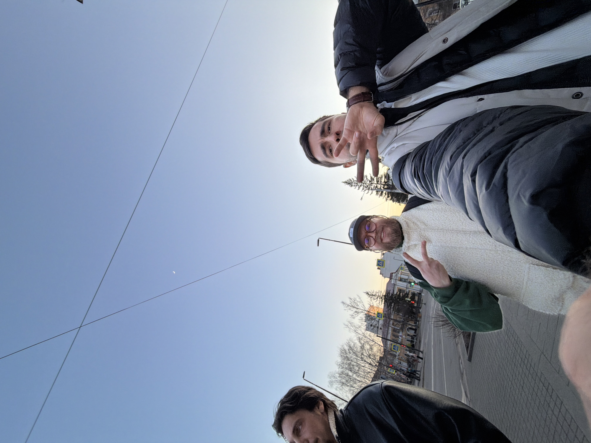 This low-angle, selfie-style photograph captures three men outdoors in what appears to be an urban setting during daylight hours. The image is taken from below, looking upwards towards the sky and the subjects.
The subject closest to the camera, partially visible in the bottom right, wears a black leather-look jacket with a white collar and looks slightly down and to the left. In the middle of the frame, a man with a beard and glasses, wearing a dark cap, a white textured vest over a green long-sleeved shirt, smiles while making a "peace" sign with his right hand. The third man, further back and positioned towards the top of the frame, wears a black puffer jacket over a white top and a brown-strapped watch. He looks directly at the camera, making a hand gesture with his left hand, possibly a "peace" sign or similar.
The background reveals a clear, light blue sky with a faint crescent moon visible in the upper right. Several thin power lines or cables crisscross the sky. Below the horizon line, an urban street scene unfolds. Bare trees line the street, indicating a cooler season. Buildings with multiple stories, featuring windows and architectural details, are visible in the distance. Streetlights and yellow diamond-shaped pedestrian crossing signs on poles can be seen along the street. A paved sidewalk is visible in the foreground and mid-ground.
The activity appears to be a casual group photo or selfie taken during an outing in the city. The sky's brightness and the visibility of the moon suggest either early morning or late afternoon/early evening. No discernible text is visible in the image.