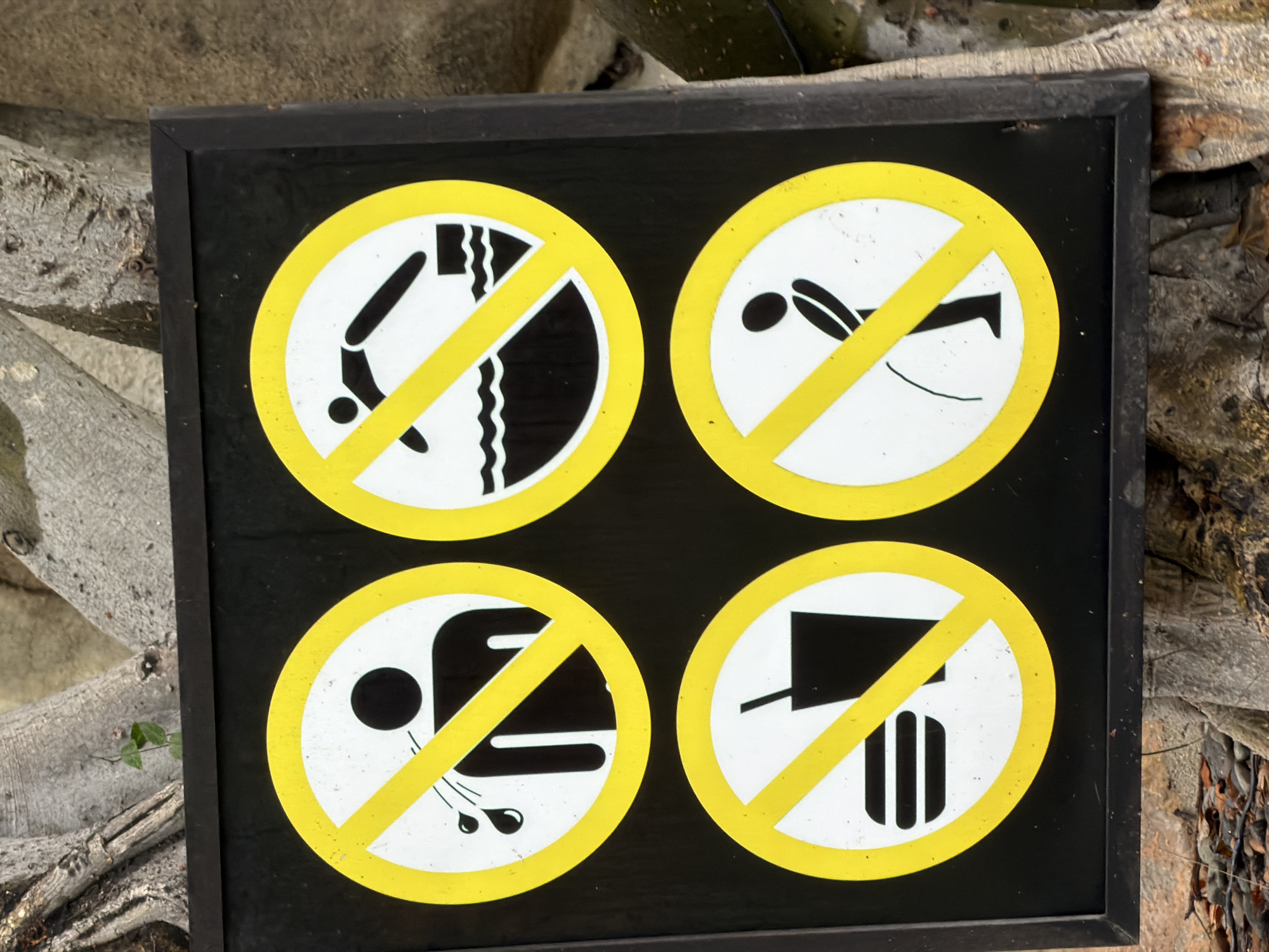The image shows a black sign with four circular prohibition symbols. Each symbol consists of a white circle with a yellow diagonal line crossing it. Inside the circles are black pictograms. The top left symbol depicts a person with water droplets coming from their mouth, indicating no spitting. The top right symbol shows a person diving into water, meaning no diving. The bottom left symbol displays a hamburger and a cup, signifying no food or drinks. The bottom right symbol illustrates a person urinating, prohibiting public urination. The sign is positioned outdoors, with large tree trunks visible in the background. There is no indication of the city of Nha Trang, Vietnam in the image.