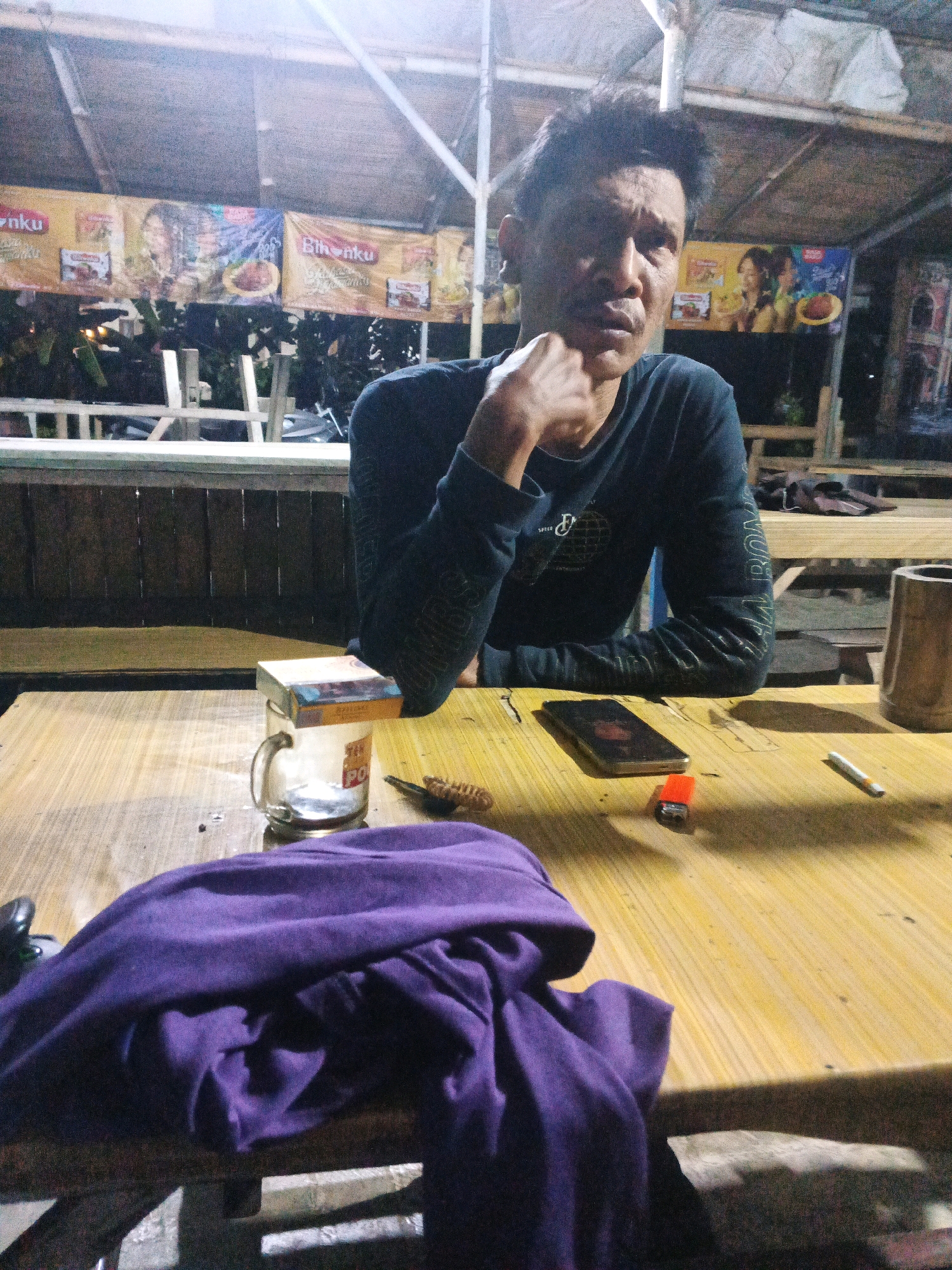 The image depicts an outdoor seating area, likely a warung or small roadside eatery, at night in Ungaran, Indonesia. A middle-aged man with dark, tousled hair and a stern expression is seated at a wooden table, resting his chin on his hand and looking directly at the camera. He is wearing a dark long-sleeved shirt. On the table in front of him are several items: a pack of cigarettes placed atop a clear glass mug, a red lighter, a single unlit cigarette, a smartphone, and a dark purple garment draped over the edge of the table. In the background, banners with text and images of food are visible, suggesting a commercial establishment. The overall atmosphere is dimly lit, with overhead lighting casting shadows and creating a somewhat gritty ambiance.