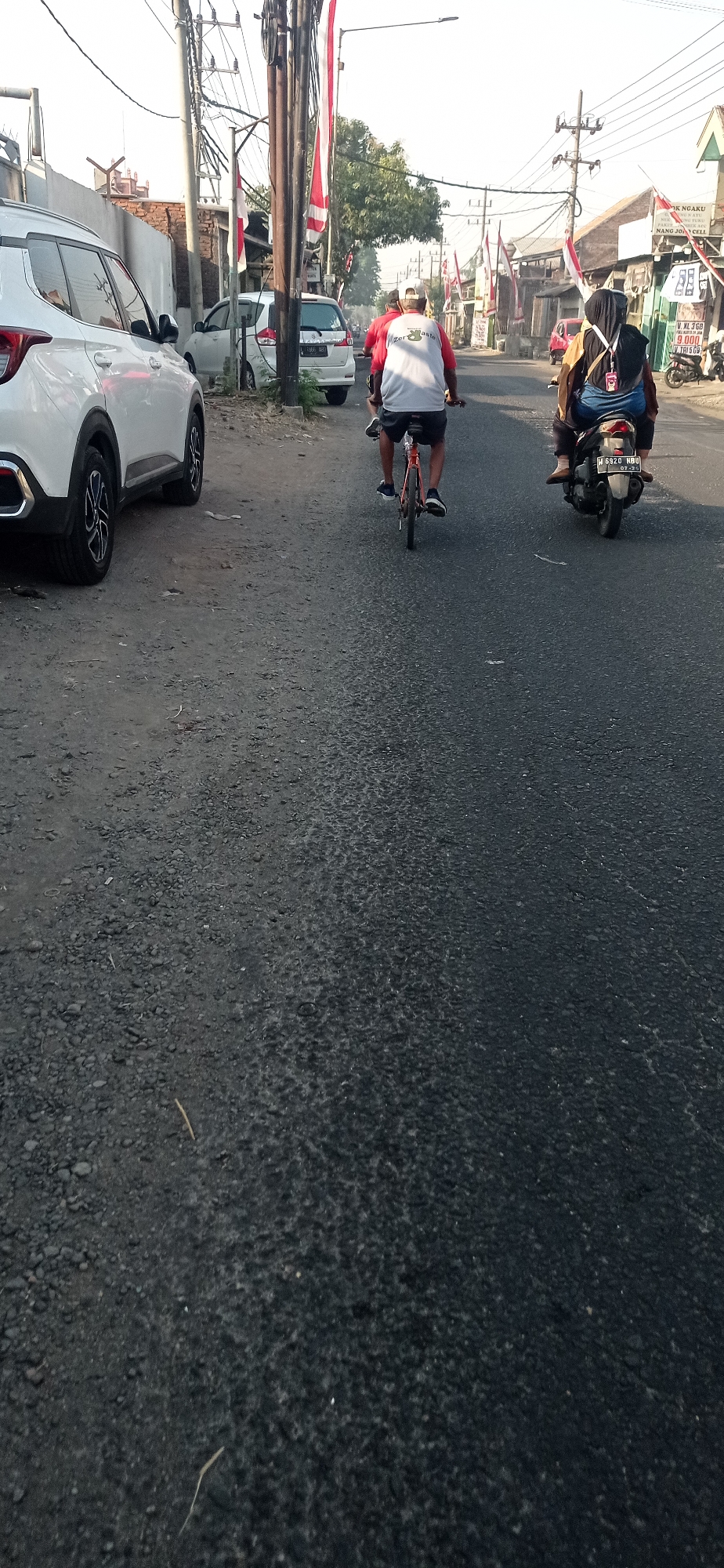 The image captures a street scene in Desa Sadang, Indonesia, likely during the daytime with moderate sunlight. On the left, a white SUV is parked along the side of a road. Further down the road, another white car is also visible. Two people are riding bicycles on the asphalt road. The first cyclist, wearing a white shirt with red sleeves and a white cap, is in the foreground and is moving away from the camera. Behind them, a person on a motorcycle, wearing a dark hijab and a yellow and black jacket, is also traveling in the same direction. The road is lined with buildings, some of which have red and white flags displayed, indicative of a national celebration or event. Utility poles with numerous wires are present along the street. The road surface appears to be asphalt with some gravel and debris. The overall atmosphere suggests a typical day in a residential or commercial area of the town.