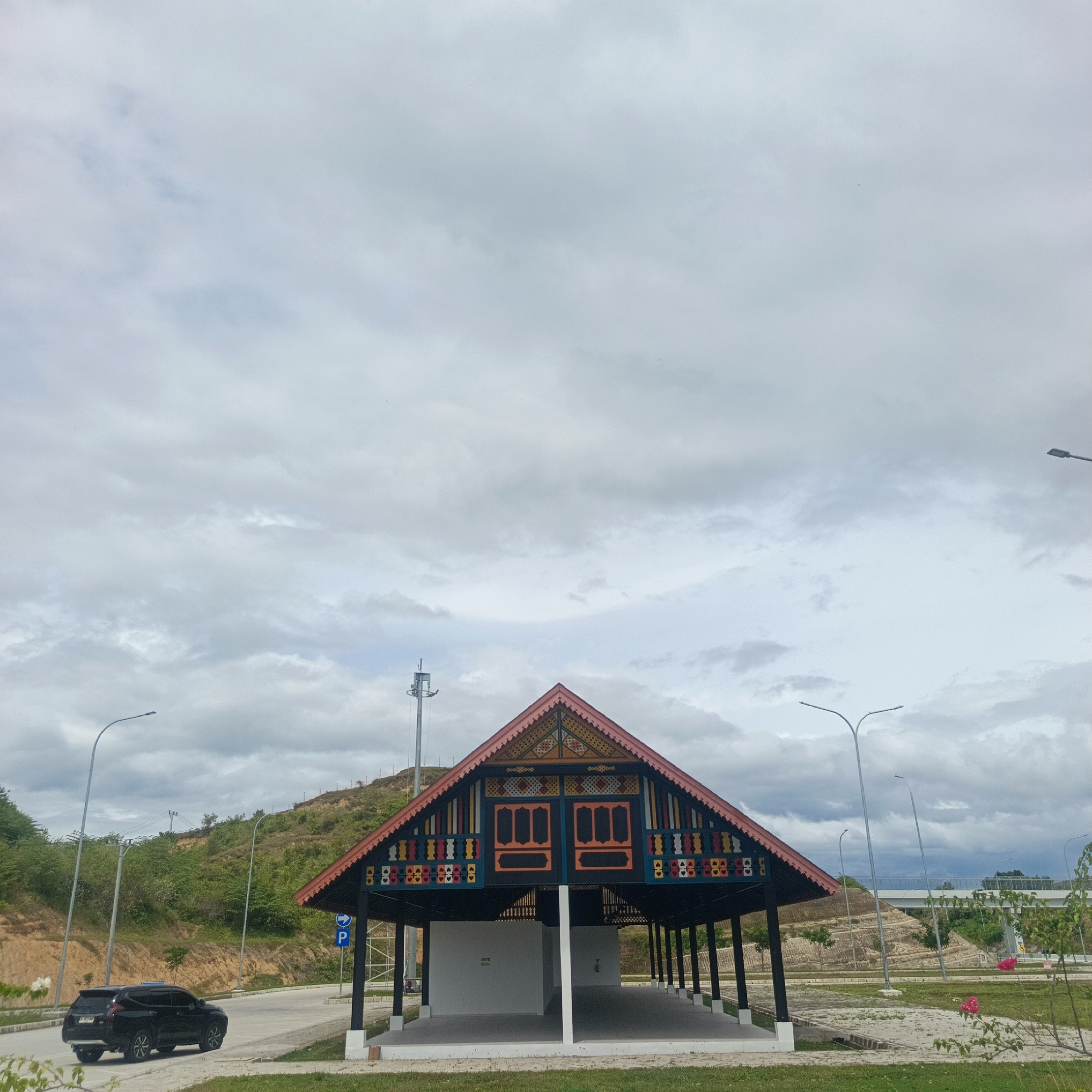 The image depicts a roadside structure with traditional Indonesian architecture in Keureuweungkrueng, Indonesia. The structure is open-sided, with a prominent, steeply pitched roof adorned with colorful geometric patterns and decorative elements in shades of red, yellow, blue, and black. The roof is supported by dark pillars. Two distinct white enclosed sections are visible beneath the roof. In the foreground, a black SUV is parked on the left side of a paved road. The environment is characterized by a slightly overcast sky, a gently sloping hillside covered with green vegetation to the left, and a modern elevated highway structure in the distance on the right. Several streetlights are visible throughout the scene, and a parking sign with a "P" and a blue arrow is posted near the structure. The ground is a mix of paved surfaces and grassy areas.