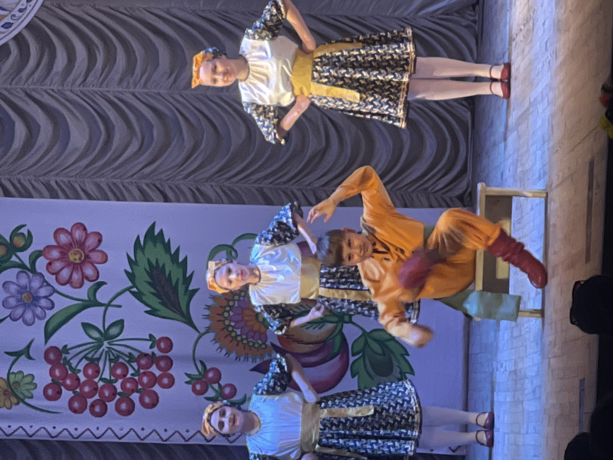 The image shows a stage performance featuring four individuals dressed in traditional folk costumes. Three young women stand on the right side of the stage, each wearing a white blouse with patterned sleeves, a yellow sash, and a dark patterned skirt. They also have yellow headscarves. A young boy is seated on a wooden bench in the center of the stage, wearing an orange tunic and red boots, with his arms outstretched. The backdrop is a large banner with a colorful floral and berry pattern on the left and dark, draped curtains on the right. The stage floor is made of light-colored wood. The scene appears to be taking place in Horbanivka, Ukraine, a village known for its cultural heritage and folk traditions, suggesting this is a local performance.