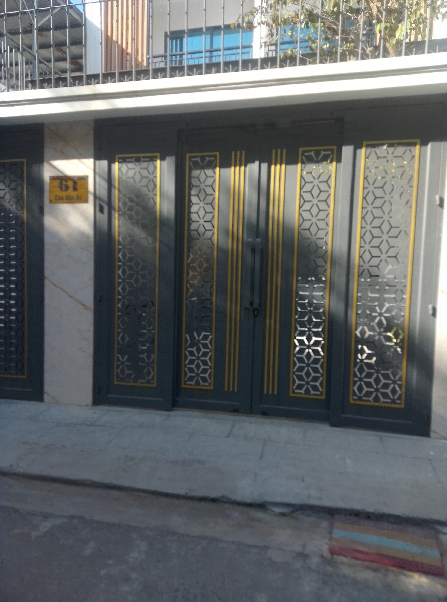 The image shows the exterior of a building, likely a residential or commercial property, located in Bac Nha Trang Ward, Vietnam. The main focus is a set of dark gray double doors with decorative yellow trim. Each door features two vertical panels of patterned cutouts, resembling a honeycomb or geometric lattice design, bordered by gold-colored framing. The doors are set within a structure that has a light-colored marbled wall on the left side, on which a yellow rectangular sign with Vietnamese text and the number "61" is affixed. The doors appear to be made of metal and have handles and a latch mechanism visible on the left door. The ground in front of the doors is paved with concrete slabs and there is a small, multi-colored welcome mat on the right side. Above the doors, a white fascia board runs horizontally. In the background, a metal railing is visible, with some greenery and a building with blue-tinted windows behind it. The overall impression is one of a well-maintained entrance to a property.