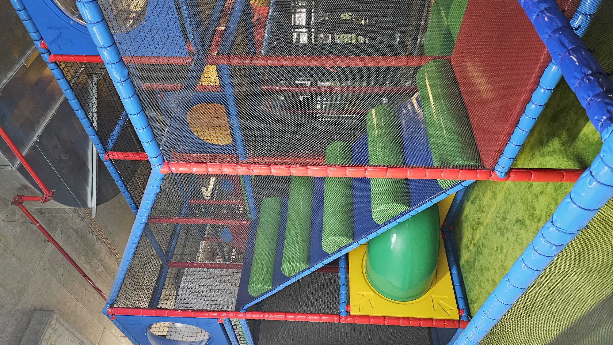 The image shows an indoor children's play structure, likely a playground or soft play area. The structure is made of brightly colored padded tubes and nets, primarily in red, blue, and green. There are several levels, interconnected with nets and padded ramps. A prominent feature is a set of stairs with green padded cylinders for steps. A yellow platform with a large green tunnel is visible at the bottom left. The floor of the play area is covered with green artificial turf. Above the play structure, exposed concrete ceiling with red pipes and metal ductwork is visible, indicating an industrial or commercial building. The scene is indoors and appears to be well-lit. The image does not provide any specific visual cues to pinpoint its location within Bratislava, Slovakia.