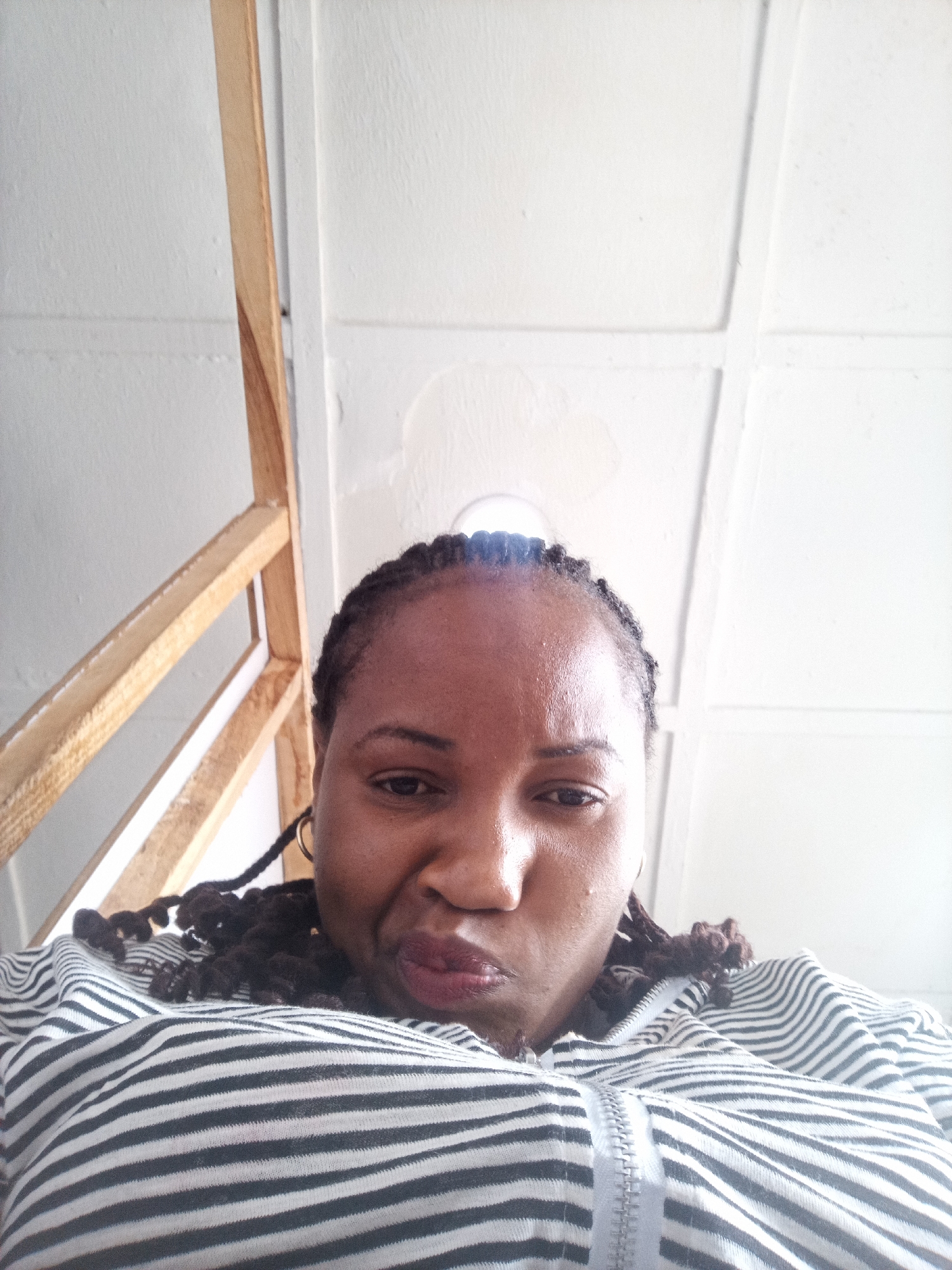 A close-up, low-angle selfie shows a woman of African descent looking directly at the camera with a distinct pout or puckered lips. Her braided hair is pulled back, and her eyes are open and engaging. She wears a long-sleeved top or hoodie with prominent horizontal black and white stripes and a visible front zipper.
The indoor background features a light-colored wooden frame structure on the left, possibly a bunk bed or shelving. The white ceiling or upper wall, which dominates the upper half of the image, displays a grid-like pattern of panels or seams. A circular bright spot, likely a light source or reflection, is visible near the top center. The scene is brightly lit. No text is visible. The image captures an informal moment indoors, potentially in Jos, Nigeria, given the provided context.