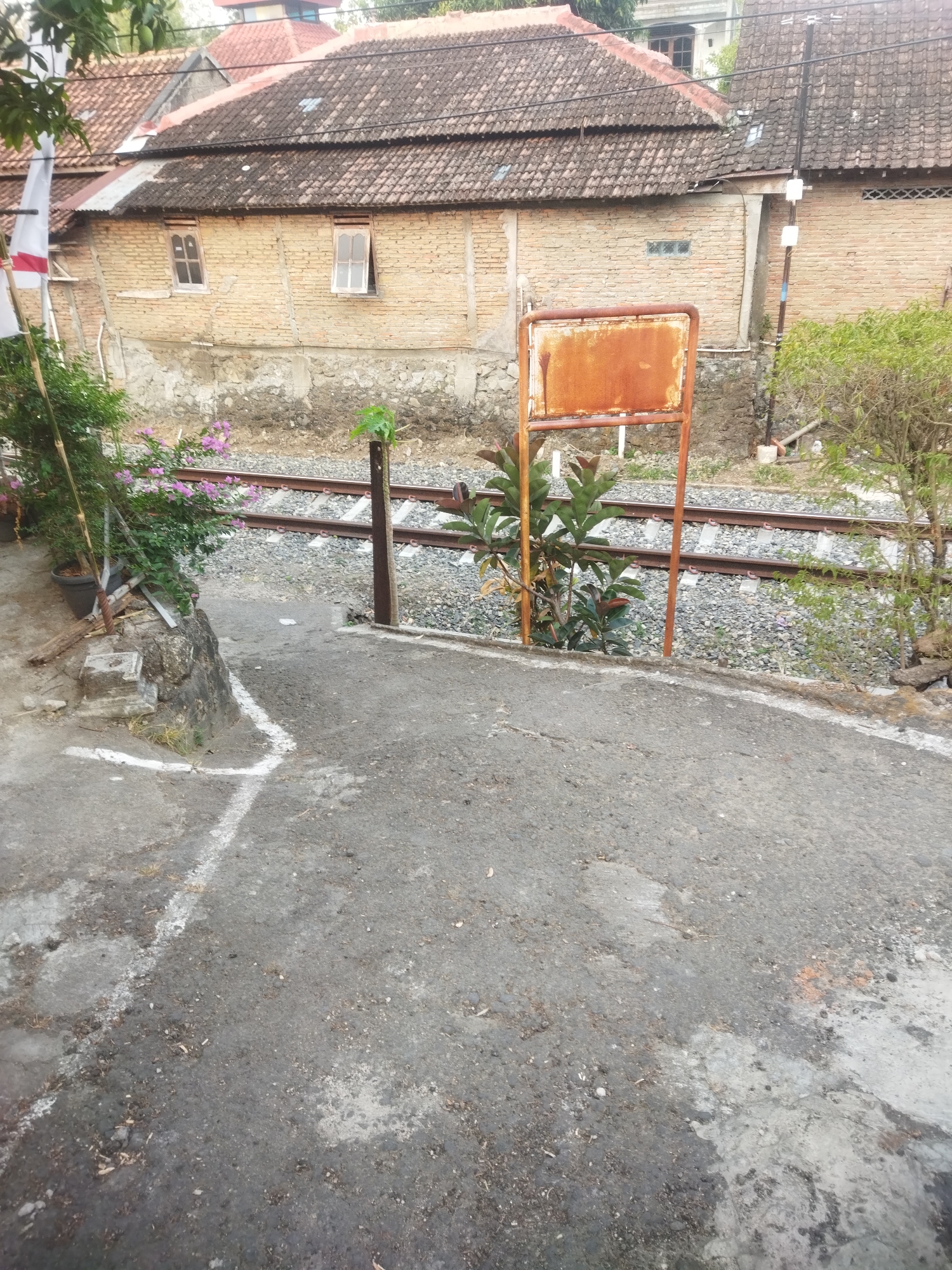 The image depicts a scene along a railway track in the city of Wonogiri, Indonesia. In the foreground, there is a paved area with some white markings, possibly indicating a pathway or lane. To the left, a small embankment with rocks and vegetation leads up to a building constructed of light-colored bricks with a tiled roof. A white and red flag is visible on a bamboo pole.
Adjacent to the embankment and in front of the brick building are train tracks laid on a bed of gravel. A rusty metal signpost with a square, weathered sign stands near the tracks, partially obscured by green plants.
Further back, the brick building stretches across the frame, featuring several windows with light-colored frames. The building has a prominent tiled roof with some sections appearing weathered. Beyond this building, other structures with tiled roofs are visible, suggesting a residential or mixed-use area. The overall environment appears to be an urban or semi-urban setting with elements of residential housing and infrastructure.