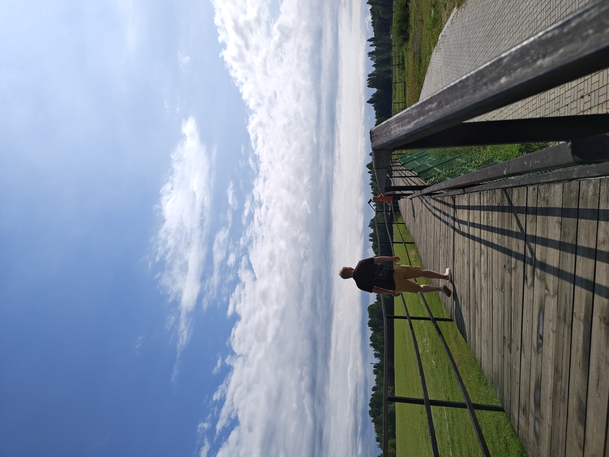 In the city of Dorishchi, Russia, a man walks along a wooden boardwalk. He is seen from behind, wearing a dark t-shirt and tan shorts. He carries a dark bag slung over his shoulder. The boardwalk is lined with a dark metal railing and a green mesh fence. Beyond the fence is a large, flat green field. In the distance, a line of trees forms the horizon under a partly cloudy blue sky. Another person, wearing an orange shirt, is visible further down the boardwalk in the distance. The boardwalk is constructed of weathered wooden planks. A paved path runs parallel to the boardwalk on the right side of the image. The sun casts long shadows across the wooden planks.