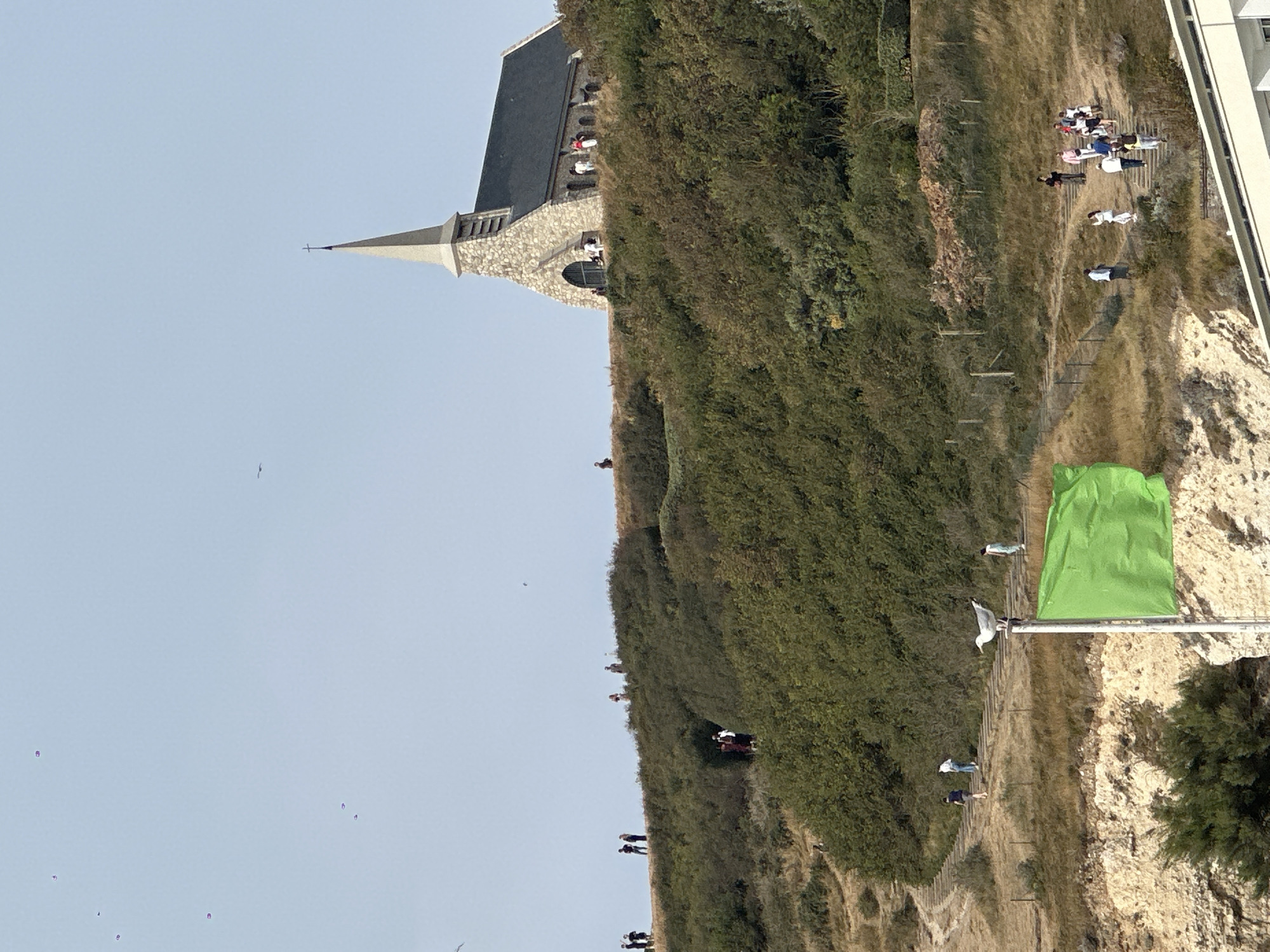 The image shows a stone chapel with a steeple situated atop a grassy hill. The chapel has a dark roof and several people are visible standing near its entrance and on the surrounding terrain. A path with steps winds up the hill, and various individuals are walking along it. In the foreground, a seagull is perched on a pole next to a bright green flag. The environment is characterized by dry grass, green bushes, and a clear sky. The scene is likely the Chapelle Notre-Dame de la Garde in Étretat, France, known for its prominent location overlooking the sea and its picturesque setting on the cliffs.