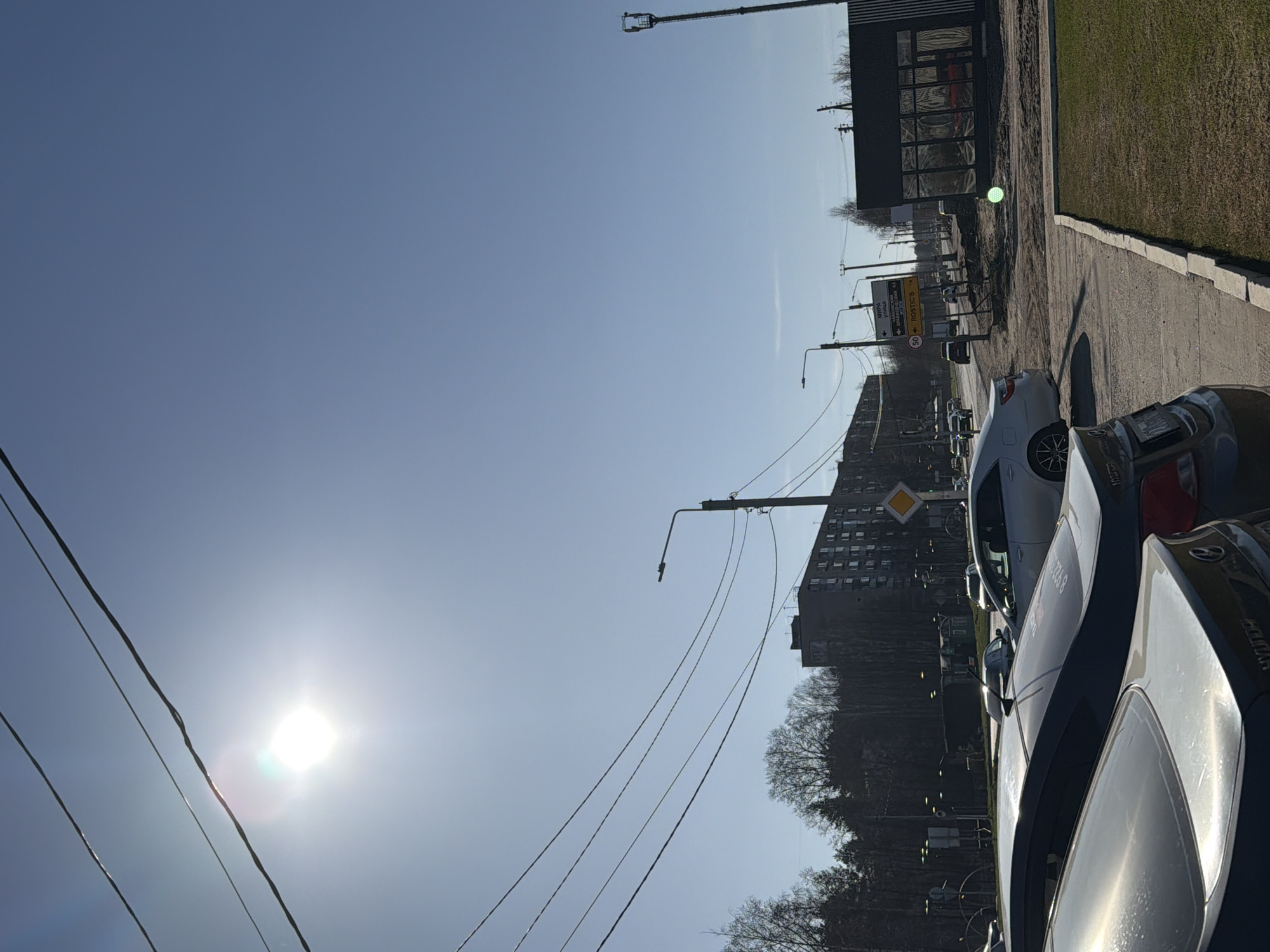 The image displays a bright, sunny street scene, captured with a 90-degree clockwise rotation, showing the sky on the left and the ground on the right. When viewed correctly, a clear, light blue sky dominates the upper portion, featuring the intensely bright sun in the lower-left, causing a prominent lens flare. Several dark power lines traverse the sky diagonally from left to right.
In the mid-ground and background, an urban environment unfolds. A paved road or parking area occupies the right side of the frame, lined by a concrete curb and a strip of green grass further right. Several cars are parked along the road; the closest one in the foreground is dark-colored, showing reflections on its roof and rear window. Another lighter-colored car is visible further down the road.
Utility poles, fitted with streetlights, punctuate the scene, supporting the power lines. Mounted on one pole are several street signs in Cyrillic script. A yellow rectangular sign points left to "МИРА УЛИЦА" (Mira Street) and right to "РОСТИКС" (Rostik's, a fast-food chain). Below it, a circular red-bordered sign indicates a "50" speed limit. Further down, a yellow diamond-shaped sign signifies a priority road.
In the background, a dark, multi-story building with numerous windows stands against the sky. To the far right, a modern-looking dark building with large, reflective windows suggests a commercial establishment. Bare trees, likely deciduous and indicating an early spring season, are scattered throughout the background and mid-ground. No people or specific activities are visible, portraying a typical, quiet daytime street in Chaikovsky, Russia. Text "8 922 602 8" is partially visible on the door of one of the cars.