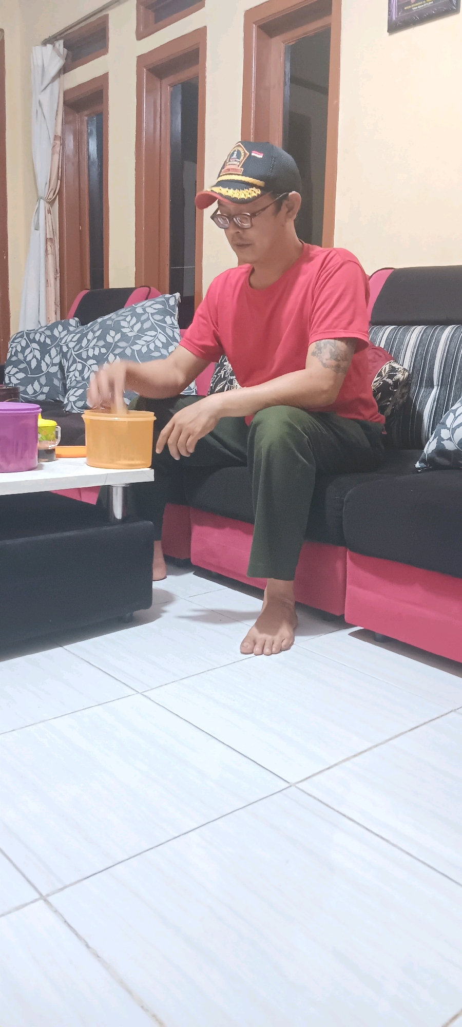 The image shows a man sitting on a couch in what appears to be a living room. The man is wearing a red t-shirt, dark green pants, and a baseball cap. He is looking down at an orange plastic container on a white table in front of him. His right hand is reaching into the container. There are two pillows with a floral pattern on the couch behind him, and another couch with black and pink upholstery to his right. To the left of the table, there is a purple container and a yellow cup with a handle. The floor is tiled with large white tiles. The background shows a wall with windows and wooden frames, and a curtain. There is a framed picture on the wall in the upper right corner. The image does not contain information that specifies the location within the city of Cingambul, Indonesia.