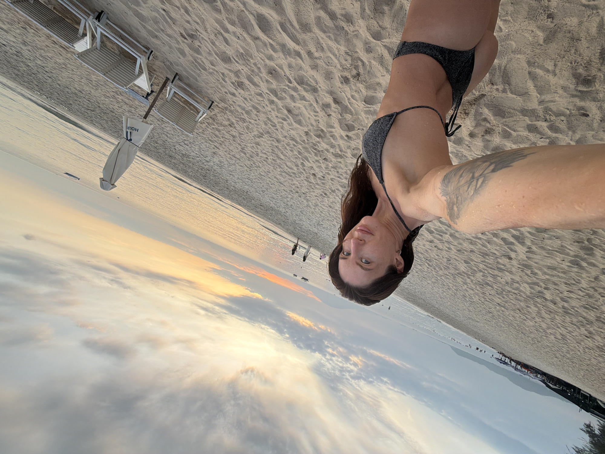 A woman wearing a sparkly black bikini stands on a sandy beach in Dong Ninh Hoa Ward, Vietnam. She is holding a phone to take a selfie, her left arm with a feather tattoo is extended. Her hair is dark brown and falls to her shoulders. The beach is wide with a gentle slope leading to the ocean. In the background, the sun is setting over the water, casting a warm glow on the clouds. There are a few lounge chairs and a closed umbrella on the sand to the right. In the distance, there are some structures on the shore and a few small boats on the water. The sky is filled with wispy clouds tinged with orange and yellow from the setting sun.