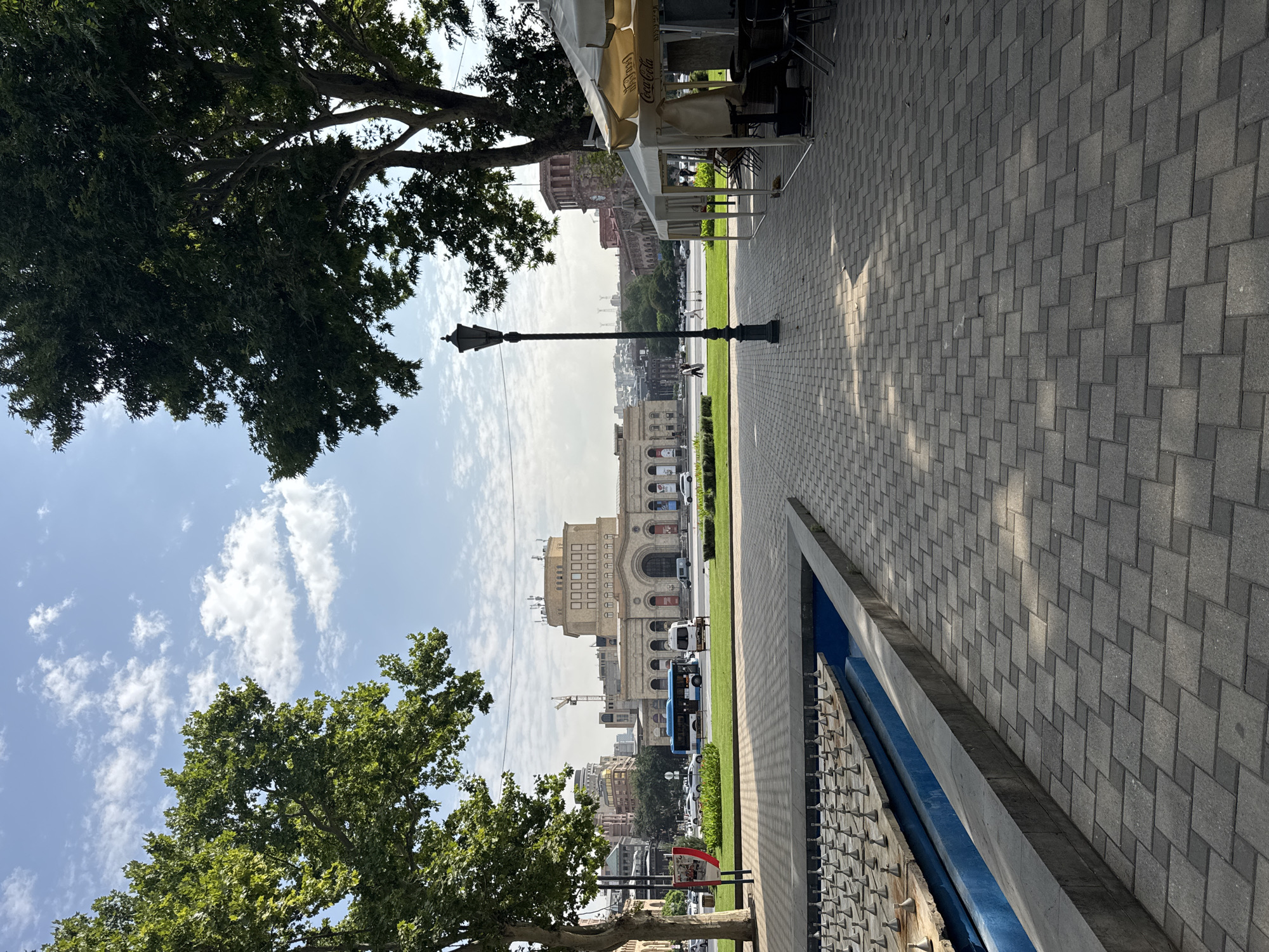 The image captures a view of Republic Square in Yerevan, Armenia. In the foreground, a paved walkway with a patterned arrangement of grey interlocking pavers leads to a partially visible, shallow blue water feature with rows of silver spray nozzles. To the right, a cafe setup is visible with white canopies, tables, and chairs, one canopy featuring a Coca-Cola logo. A tall, ornate black lamppost stands prominently in the middle ground, casting a shadow on the walkway. Beyond the walkway and a strip of green grass, a street with traffic, including a blue bus and white van, is visible. In the background, the grand architecture of the National Gallery and History Museum of Armenia dominates the scene, characterized by its neoclassical style with arched windows and ornate facade. The sky above is blue with scattered white clouds. Two large, leafy trees frame the view on the left and right, with their branches reaching towards the sky. In the distance, other buildings and a construction crane are visible, indicating the urban environment of Yerevan.