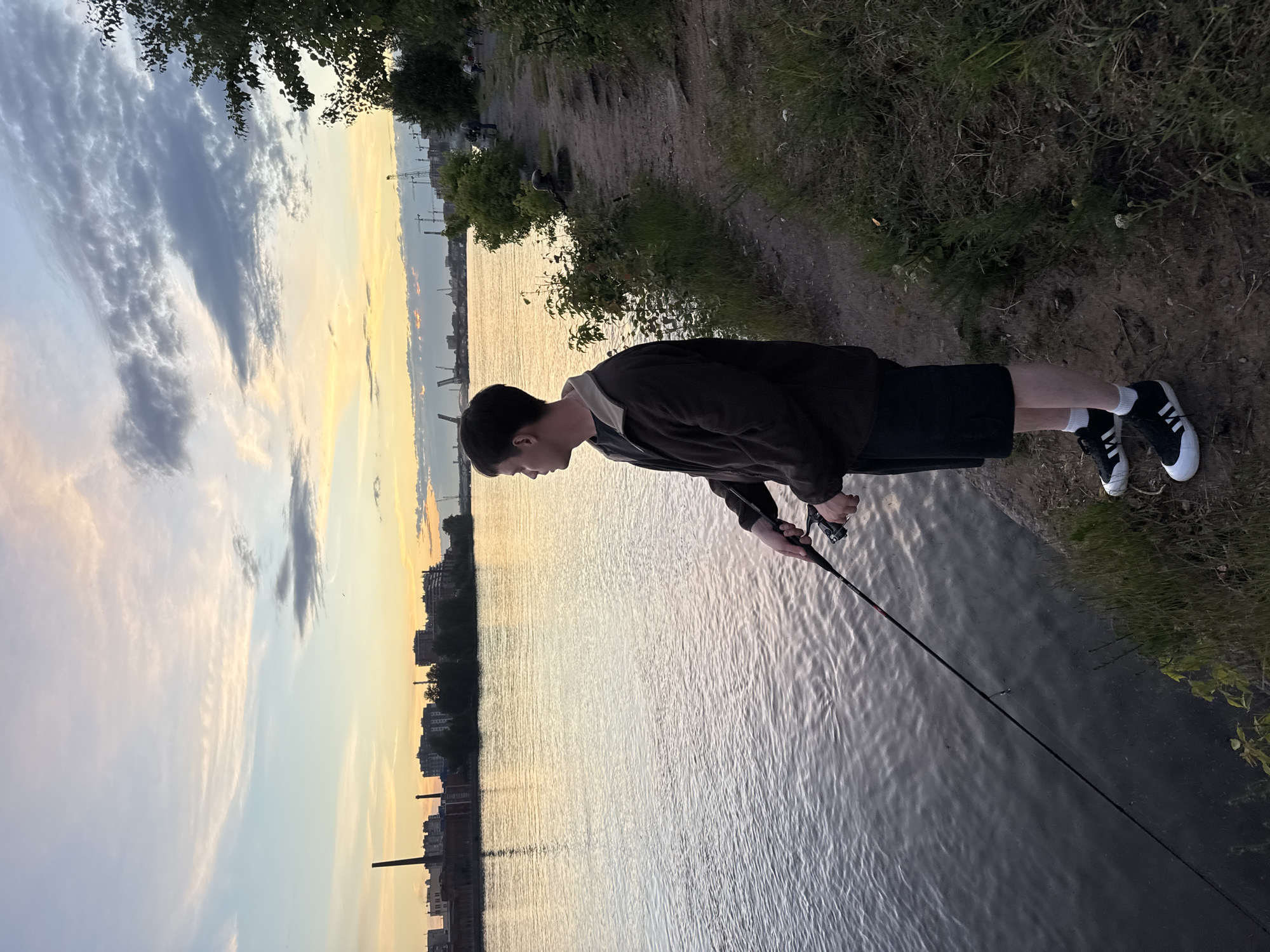 In Saint Petersburg, Russia, at sunset, a young man stands on the bank of the Neva River, fishing. He is dressed in a dark brown jacket, black shorts, white socks, and black and white sneakers. He holds a fishing rod with a reel, casting his line into the water. The river reflects the soft hues of the sky, a blend of pale blue, pink, and orange, with scattered clouds. In the distance, across the river, is the industrial silhouette of the city, with buildings and cranes visible. To the right of the man, a dirt path lined with grass and trees stretches along the riverbank, where other people can be seen in the distance. The overall scene depicts a peaceful evening activity by the water.