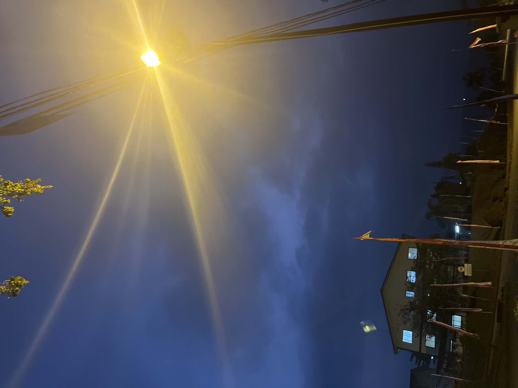 The image depicts an outdoor scene at dusk or night, with a bright streetlamp dominating the right side of the frame, casting a strong yellow glow. The sky is a deep blue, with wispy clouds visible. In the background, a two-story building with illuminated windows is seen, surrounded by trees and several flags on poles, some of which appear to be Indonesian flags with red and white stripes. The lighting suggests artificial illumination from the streetlamp and the building. The location is identified as Lalampu, Indonesia. There are no people visible in the image.