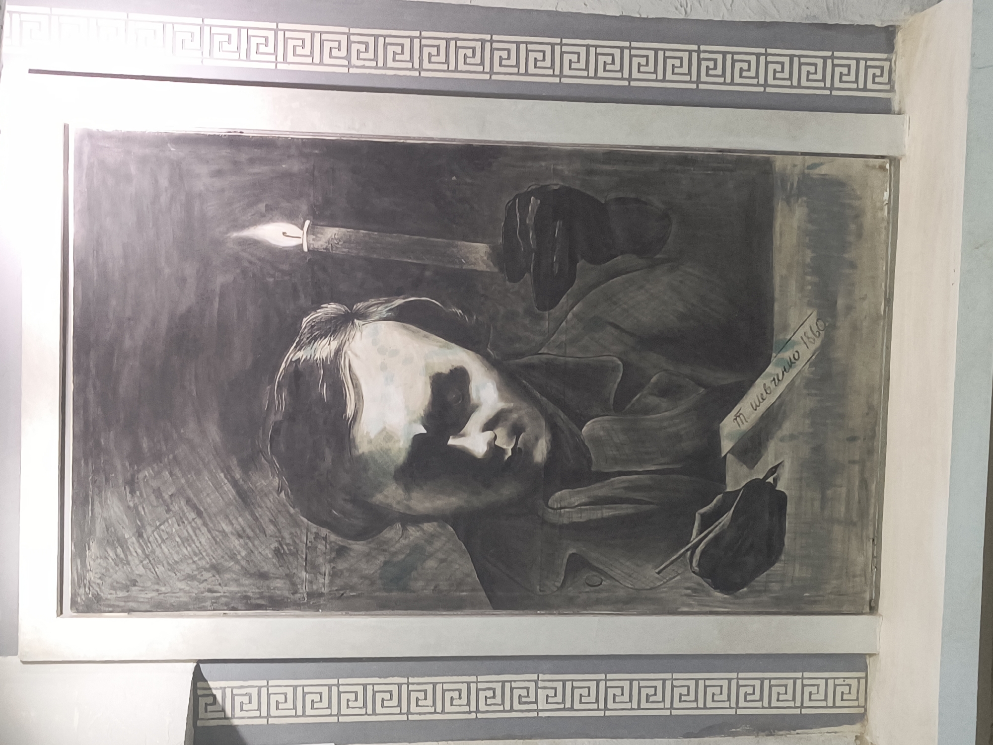 The image displays a black and white mural depicting a man writing by candlelight. The man is shown in profile, with his head tilted downwards as he focuses on his writing. He is holding a quill pen in his right hand and a lit candle in his left. The candlelight illuminates his face, casting shadows and highlighting his features. The mural is framed by a decorative border featuring a Greek key pattern. The overall style of the artwork is somber and dramatic, emphasizing the act of creation and contemplation. This mural is located on the wall of a building in Rivne, Ukraine.