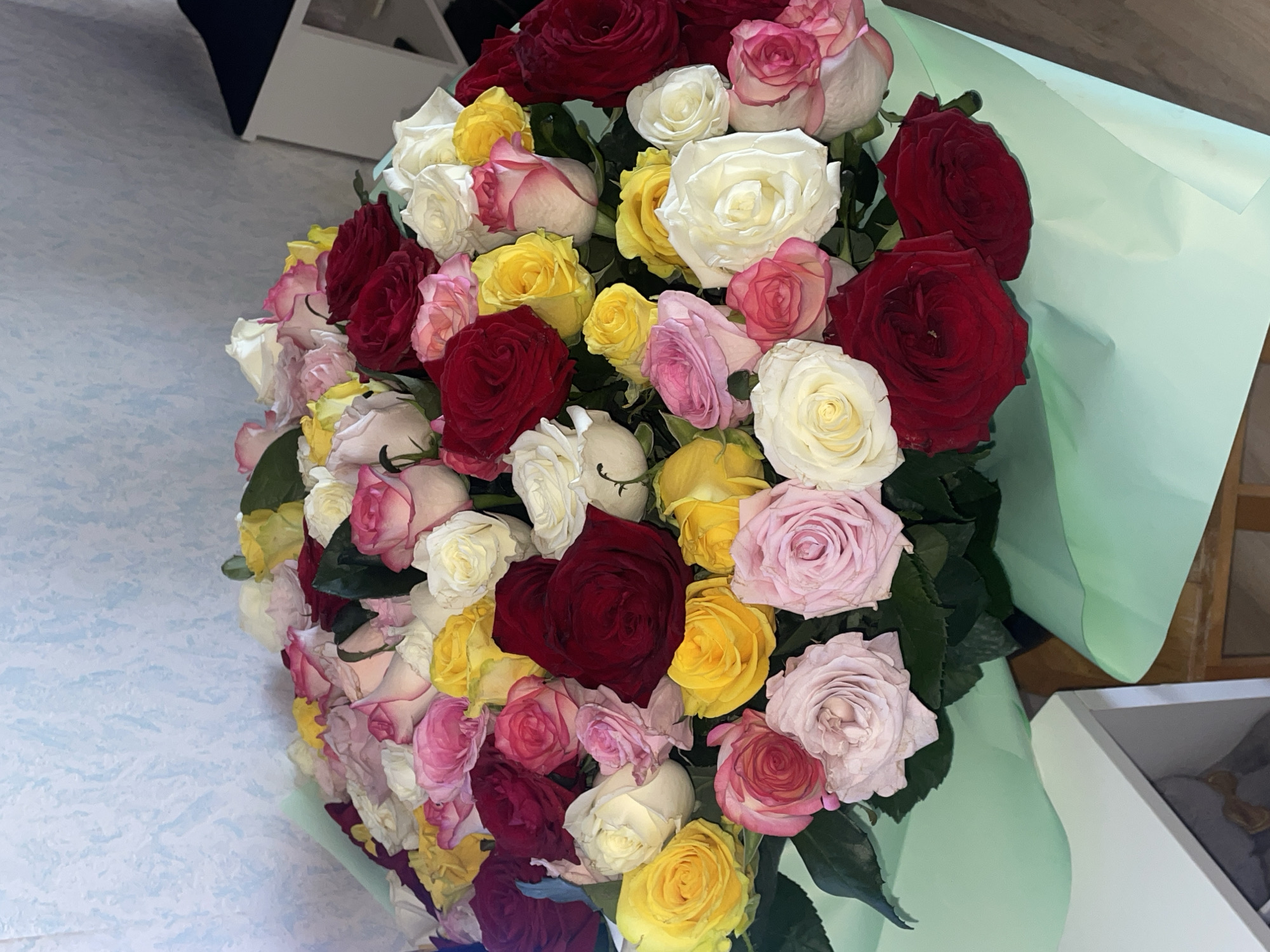 This media file captures a large, dense bouquet of roses, presented from a slightly elevated angle. The bouquet features a rich assortment of roses in various colors, including deep red, bright yellow, pure white, and diverse shades of pink, ranging from pale blush to vibrant fuchsia. Green foliage is visible among the blooms, enhancing the natural appeal. The entire floral arrangement is wrapped in light green or mint-colored floral paper, which frames the bouquet on the right and bottom.
The background on the left side of the image shows a light-colored surface with a subtle, textured pattern, possibly a floor or tablecloth. In the upper left corner, parts of a dark object and a white, angular structure (likely furniture or shelving) are visible. Another white object, possibly a box, can be seen in the lower right background.
No individuals are present, and no specific activity is occurring. However, the presence of such a bouquet typically signifies a celebratory occasion, a gift, or an expression of affection. The vibrant mix of colors often conveys sentiments of joy, love, and friendship. The scene is indoors, and there is no visible text. The location context is Votkinsk, Russia.