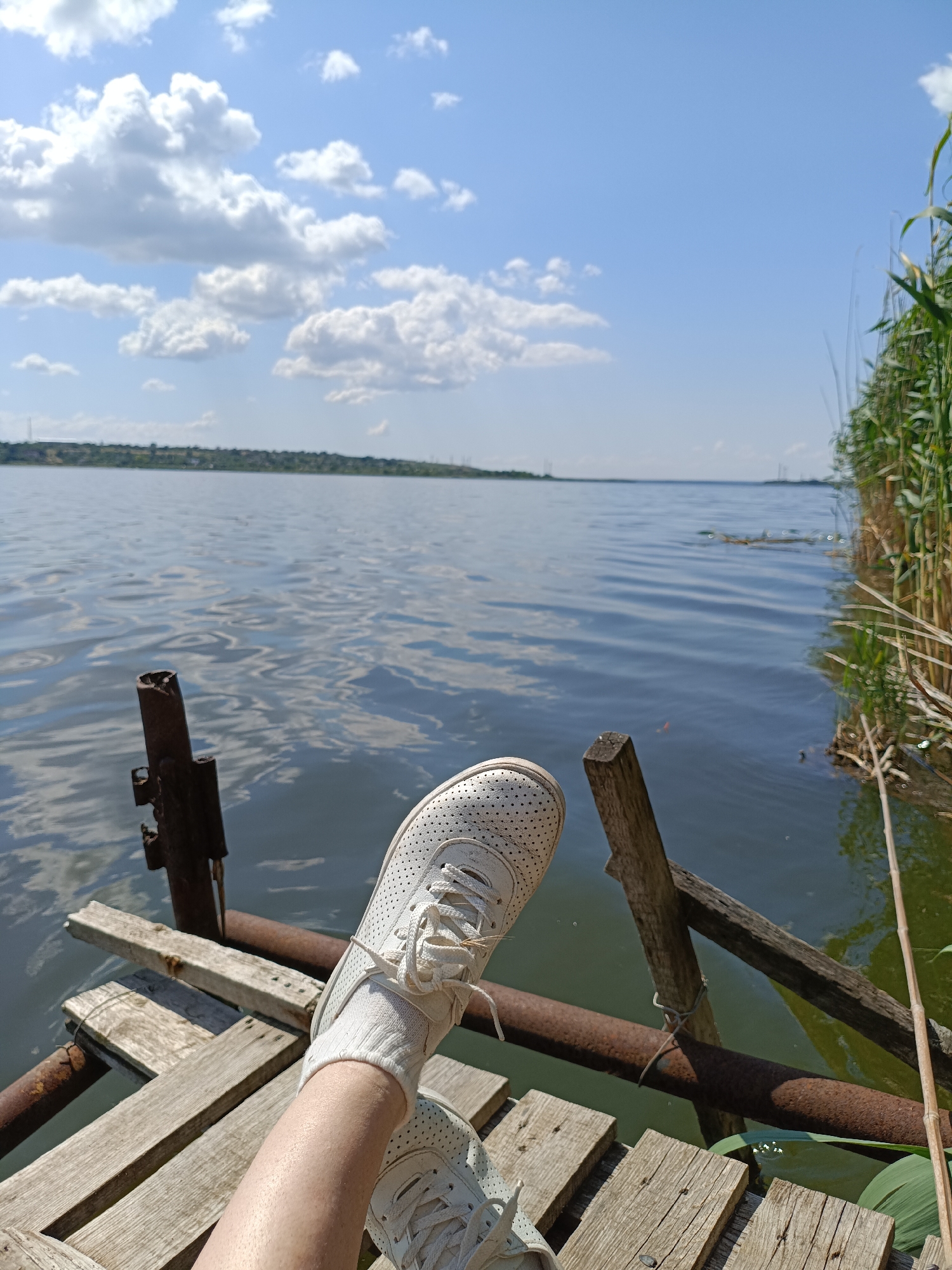 A person's legs, clad in white sneakers and white socks, are positioned on a wooden pier over a body of water. The pier is constructed of weathered wooden planks and supported by rusty metal pipes and wooden posts. The water is calm with ripples reflecting the blue sky and scattered white clouds. Tall reeds grow along the right bank of the water. In the distance, a hazy shoreline with a few scattered buildings and power lines is visible under a clear blue sky. The scene appears to be set in a natural outdoor environment.