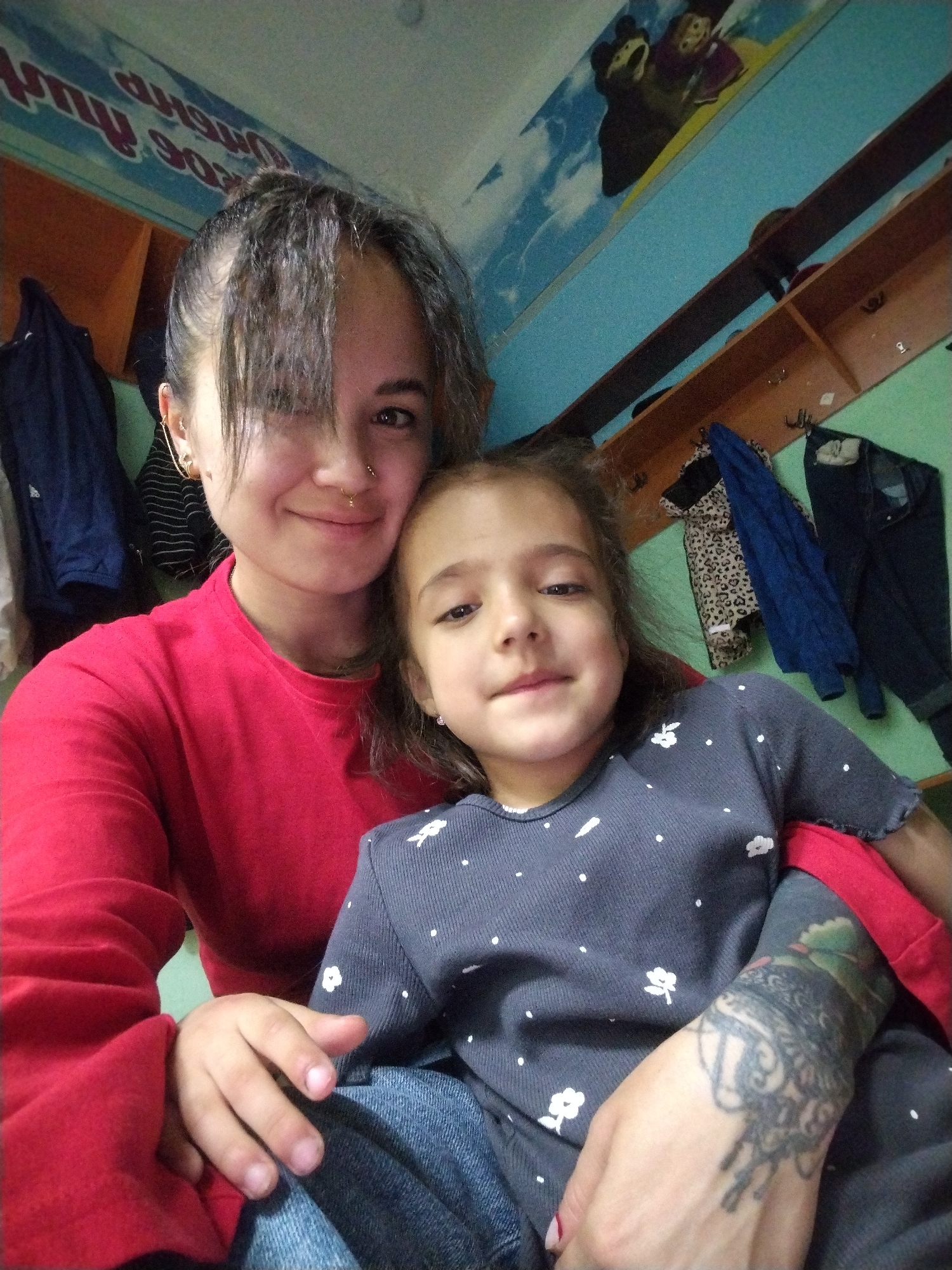 This is a selfie taken indoors, likely in a child's room given the cartoon mural on the wall. The subjects are two people: an adult woman and a young girl. The woman, on the left, has dark hair pulled back with some bangs falling forward, and is wearing a bright red long-sleeved shirt. She has a nose ring and ear piercings. The young girl, on the right, has dark hair and is wearing a dark grey shirt with a white floral pattern. She is nestled in the woman's arms, and the woman's tattooed arm is visible around the girl. The background includes shelves with clothes hanging on them and a section of a wall with a mural depicting cartoon characters. The lighting suggests it is daytime. The mood appears affectionate and close. The text on the wall, possibly a sign or decoration, is partially visible and appears to be in Russian, with some characters resembling Cyrillic letters and numbers like "903".