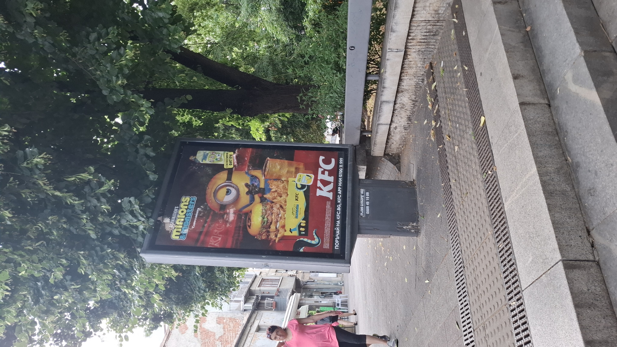 A man in a pink shirt and black shorts is lying on the ground near a billboard. The billboard is for KFC and features a Minions character, a burger, and fried chicken. Text on the billboard reads "KFC", "MINIONS & MONSTERS", and "ПОРЪЧАЙ НА KFC.BG, KFC APP ИЛИ 0700 11 999". Below the billboard, text reads "ДР.КИР БОРОДЖ ООД" and "0888 49 19 09". The scene appears to be outdoors during the day, with trees and a sidewalk visible. The ground is paved with stones and metal grates. Steps are visible to the right of the billboard. The overall impression is a casual urban setting.