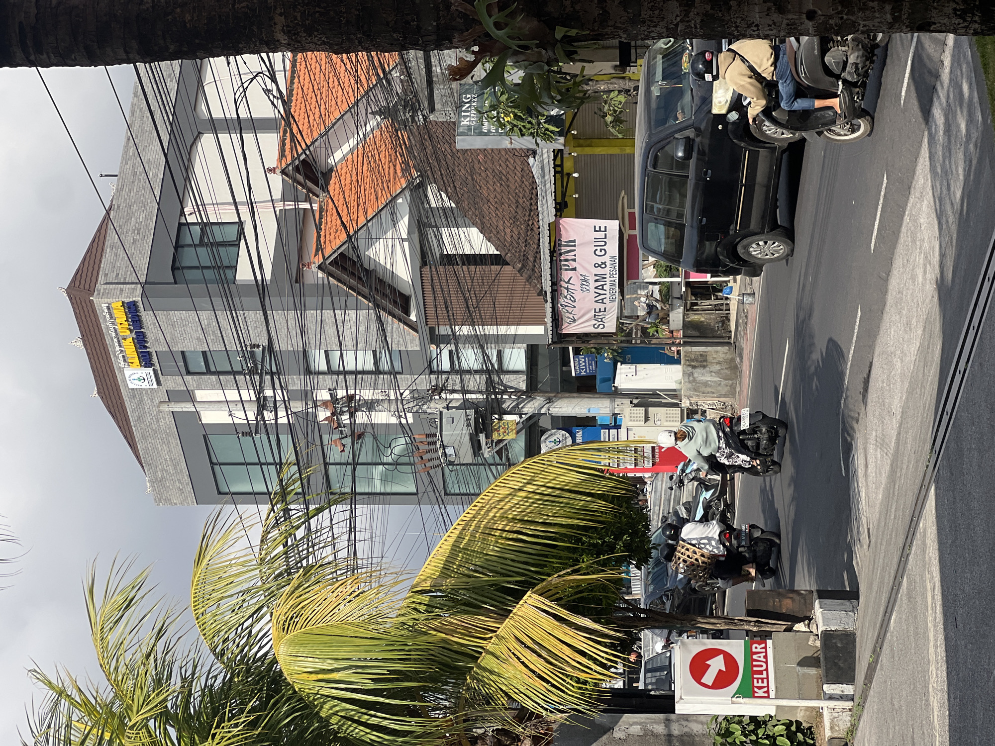 A daytime, street-level shot captures a scene in Denpasar, Indonesia, characterized by a busy roadway and multi-story buildings. The foreground features a prominent white sign with a red arrow pointing to the right and the word "KELUAR" in red, indicating an exit. To the left, a portion of a palm tree with green and yellow fronds partially obscures the view.
The street itself is paved and marked with white lane dividers. Several motorcycles and a black van are present. On the street, two individuals are riding motorcycles; one is carrying a woven basket on their back, and the other is wearing a light brown hooded jacket and jeans. A black van is also on the road.
In the background, a modern, gray, multi-story building with numerous windows is visible, adorned with a sign that reads "KLINIK UTAMA" and "SAU PURI MEDIKA." Adjacent to it, another building with a reddish-brown tiled roof is present. Various electrical wires crisscross the scene, typical of an urban environment. A pink banner with Indonesian text, "KRUSUK PINK SEDIA SATE AYAM & GULE MENERIMA PESANAN," is displayed on the lower part of the buildings. Another sign for "KING GEPERZZ" is also visible. The sky is overcast with a pale blue hue.