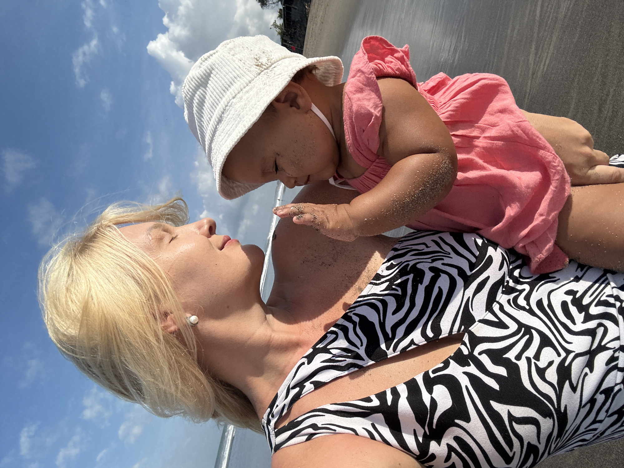 A blonde woman with her eyes closed holds a baby on a beach. The woman wears a black and white zebra-print swimsuit and a pearl earring. The baby wears a white bucket hat and a pink dress. Both are covered in sand. The background shows the ocean and a blue sky with clouds. The location is the beach in North Kuta, Indonesia.