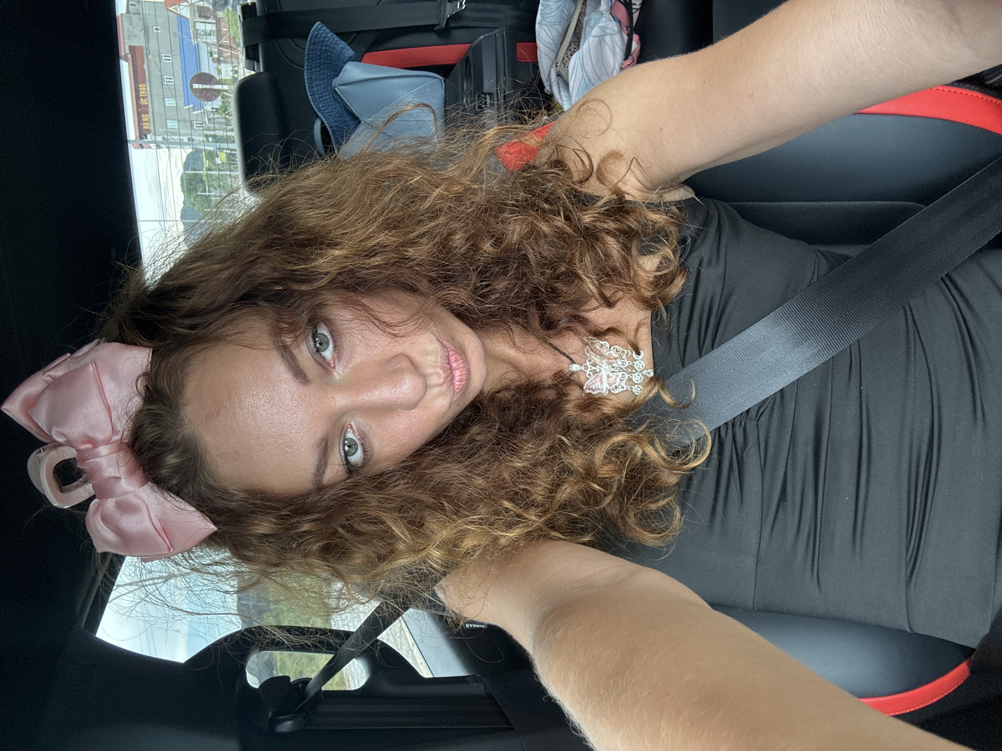 A woman with long, curly brown hair and a large pink bow on her head is taking a selfie in the passenger seat of a car. She is wearing a black, strapless, ruched dress and a silver necklace with a large, ornate pendant. Her eyes are open and she is looking at the camera with her lips pursed. The car's interior is visible, including a black seatbelt across her chest. In the background, through the car window, a building with Vietnamese writing is visible, suggesting the location is Hoa Lư Ward, Vietnam. Other car seats and luggage are also visible.