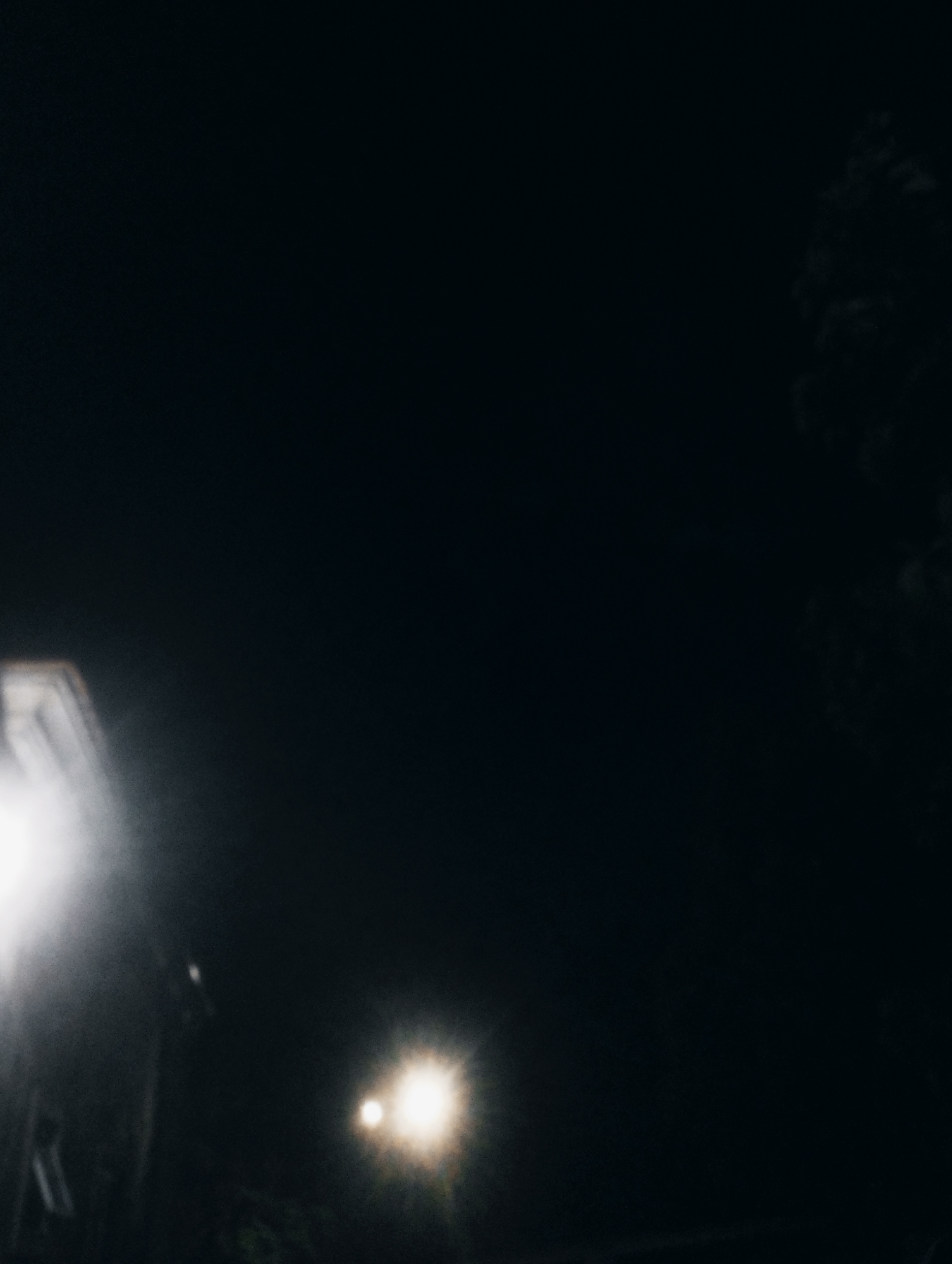 The image depicts a nighttime scene in a dimly lit area. In the foreground, on the left side, a bright light source emanates from what appears to be a structure or object, casting a strong glare. Further to the right and slightly lower, another, smaller light source is visible, also glowing brightly. The background is predominantly dark, with a faint suggestion of tree or foliage on the far right. The overall impression is that of an outdoor urban environment at night, potentially a street or an open area, illuminated by artificial lights. No specific landmarks or context related to Jos, Nigeria are discernible from the visual information provided.