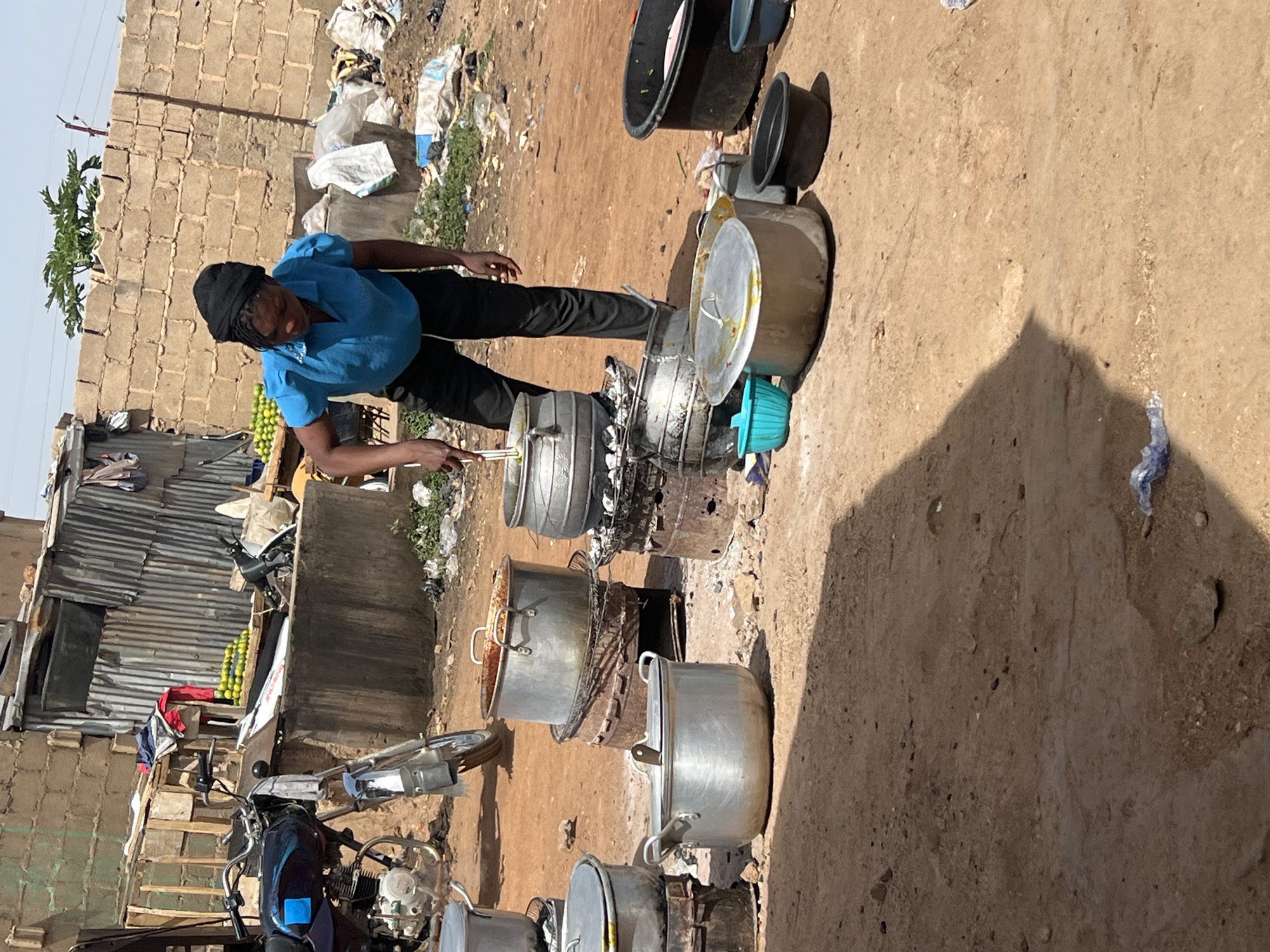 This image depicts an outdoor cooking scene in Gura Top, Nigeria, under bright, sunny conditions, likely midday or early afternoon given the shadows. A man, dressed in a blue short-sleeved shirt, black trousers, and a black head covering, is the central figure. He is actively engaged in stirring the contents of a large, aluminum cooking pot with a long utensil.
Around him, numerous other large aluminum pots, some with lids, are set up on what appear to be traditional outdoor stoves or open fires, indicated by their dark, sooty bases. These multiple large pots suggest the preparation of food on a significant scale, possibly for a community, a market, or street food sales. Several black and blue basins or buckets are also visible near the cooking area.
The ground is dry, reddish-brown dirt. In the background, there's a structure made of unrendered cinder blocks and a corrugated metal building. Next to the corrugated structure, several bags filled with bright green, spherical fruits, possibly limes or small oranges, are stacked. A motorbike is partially visible in the foreground to the left. The surroundings show signs of a bustling, utilitarian environment with some scattered debris. Visible text on a white object near the corrugated metal structure reads "ONE ZONE." The man's expression is focused on his task.