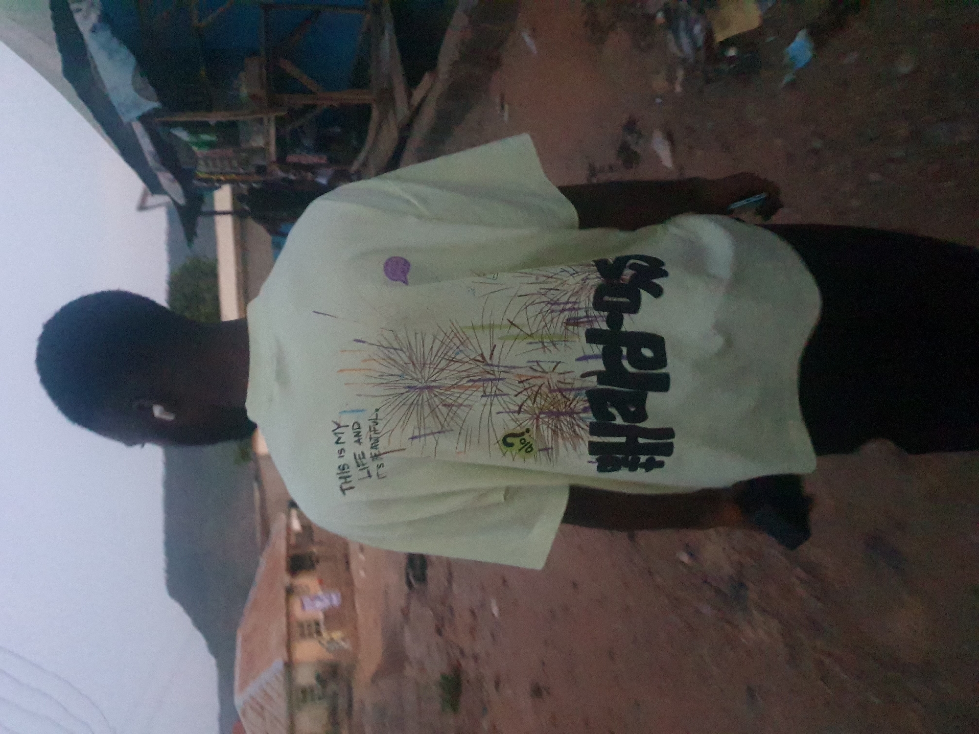 A person is wearing a light yellow shirt with black text and a firework graphic on the back. The text includes the phrase "THIS IS MY LIFE AND IT'S BEAUTIFUL". The person is holding a phone in their right hand. The background shows an outdoor environment in Lokoja, Nigeria, with buildings and possibly a mountain in the distance. The lighting suggests it might be dusk or dawn. The ground is visible.