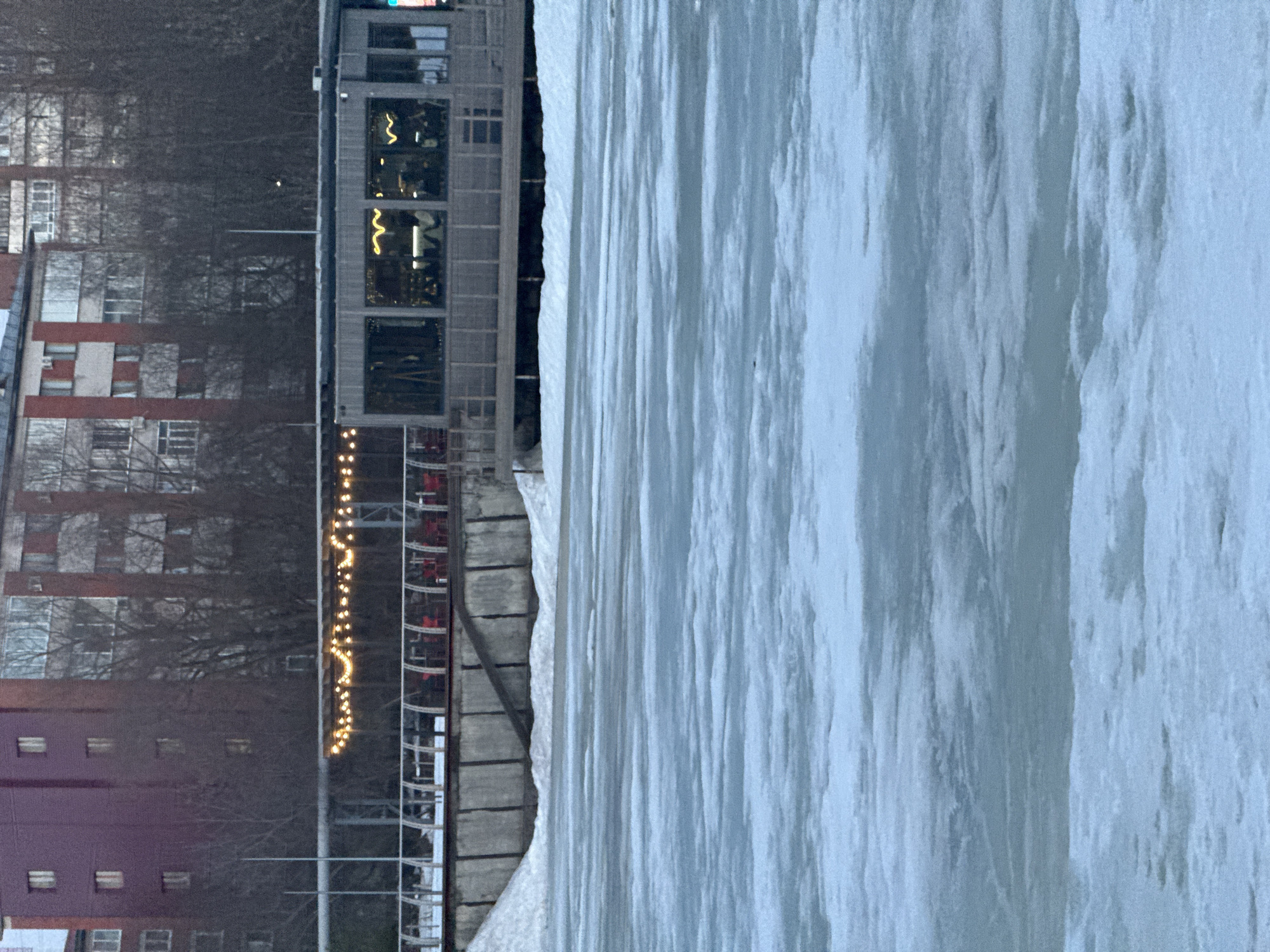 The image depicts a frozen body of water, possibly a river or lake, adjacent to buildings in Votkinsk, Russia. On the left are apartment buildings of varying colors and heights. A modern building with lit windows and strings of decorative lights is visible. The right side of the frame is dominated by a large expanse of frozen water, showing patterns and textures indicative of ice.
