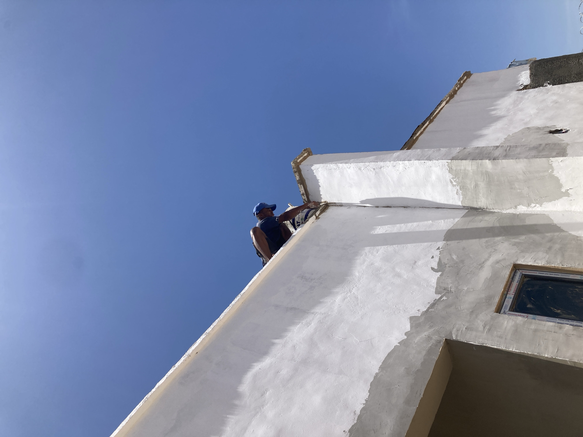 A low-angle, long shot captures a man applying stucco to the exterior of a white building against a clear blue sky. The man, wearing a blue baseball cap and dark tank top, is positioned near the top of the building, appearing to work along the edge where the roof meets the wall. The building's surface shows varying textures, with some areas covered in fresh, light-grey stucco, while others are painted white. A window frame is visible on the side of the building. The location is presumed to be in Jos, Nigeria, although no landmarks directly confirm this.