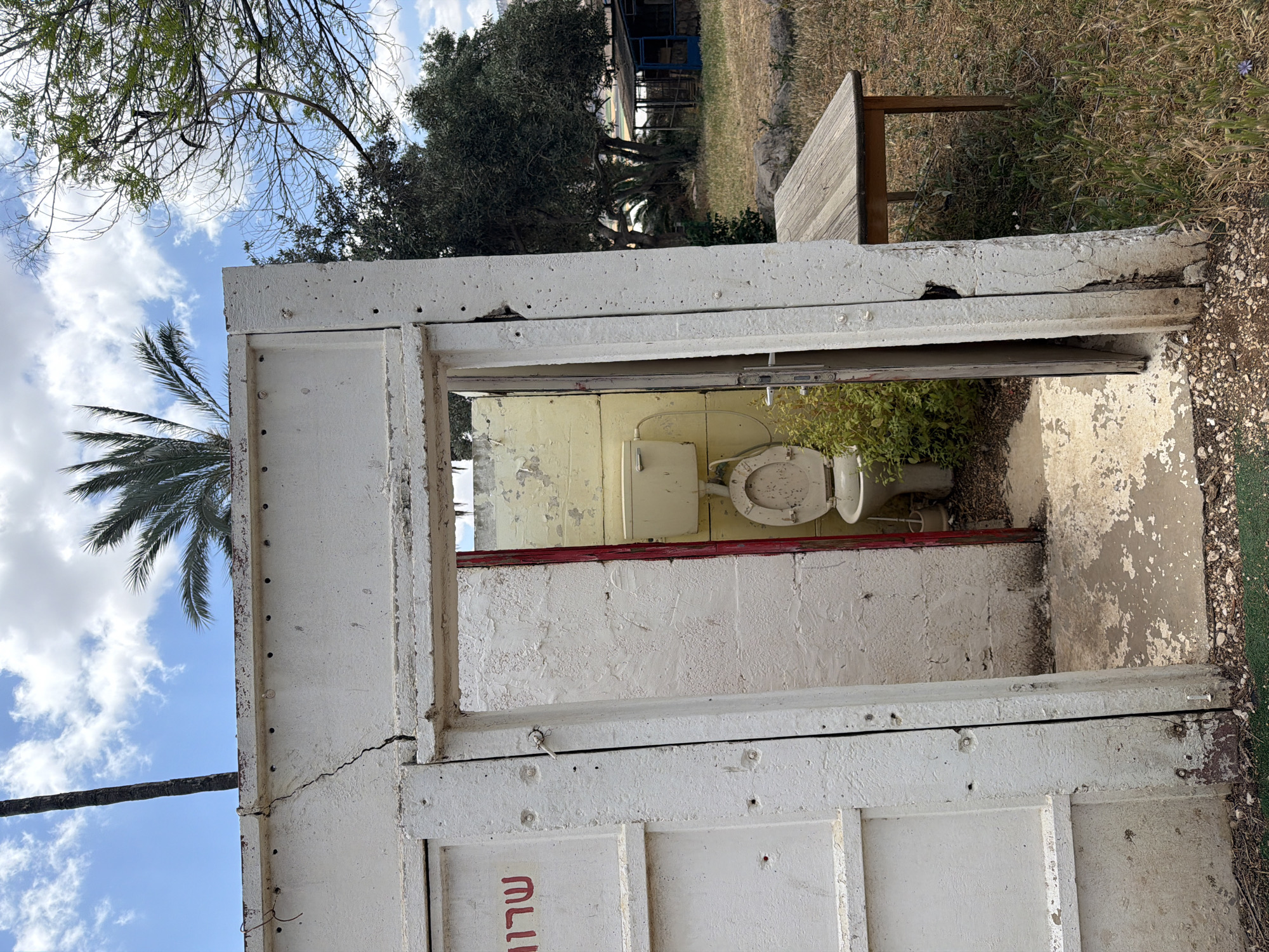 The image shows a dilapidated, outdoor toilet facility in Gilboa Regional Council, Israel. The structure is made of white-painted concrete panels, appearing worn and cracked. The Hebrew word "שירותים" (meaning "toilets") is visible on one of the panels. Inside, the toilet is yellowed and aged, partly obscured by overgrown vegetation. The surrounding environment consists of grass, trees, and a blue sky with scattered clouds. A wooden bench is located nearby.