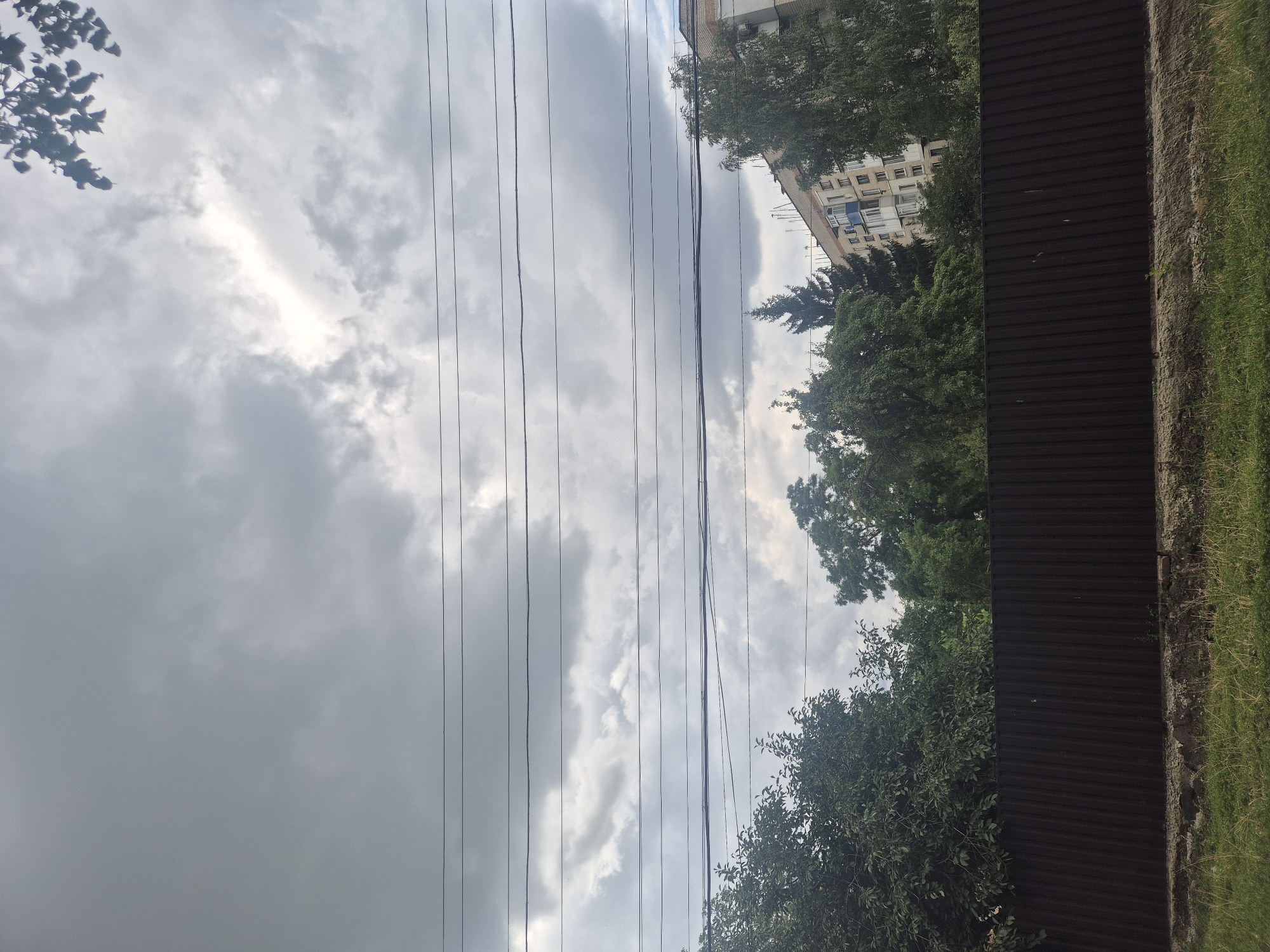 The image depicts an outdoor scene with a cloudy sky dominating the upper portion. Several parallel electrical wires are strung horizontally across the sky. Below the wires, a dark brown, corrugated metal fence runs horizontally, separating the foreground from the background. Behind the fence, lush green trees are visible. To the right of the trees, a multi-story building with light-colored walls and visible windows is partially obscured. The lower foreground consists of a strip of dry grass and some dirt. The scene appears to be in a residential area, possibly in the city of Sozonivka, Ukraine. There are no people or animals visible in the image.
