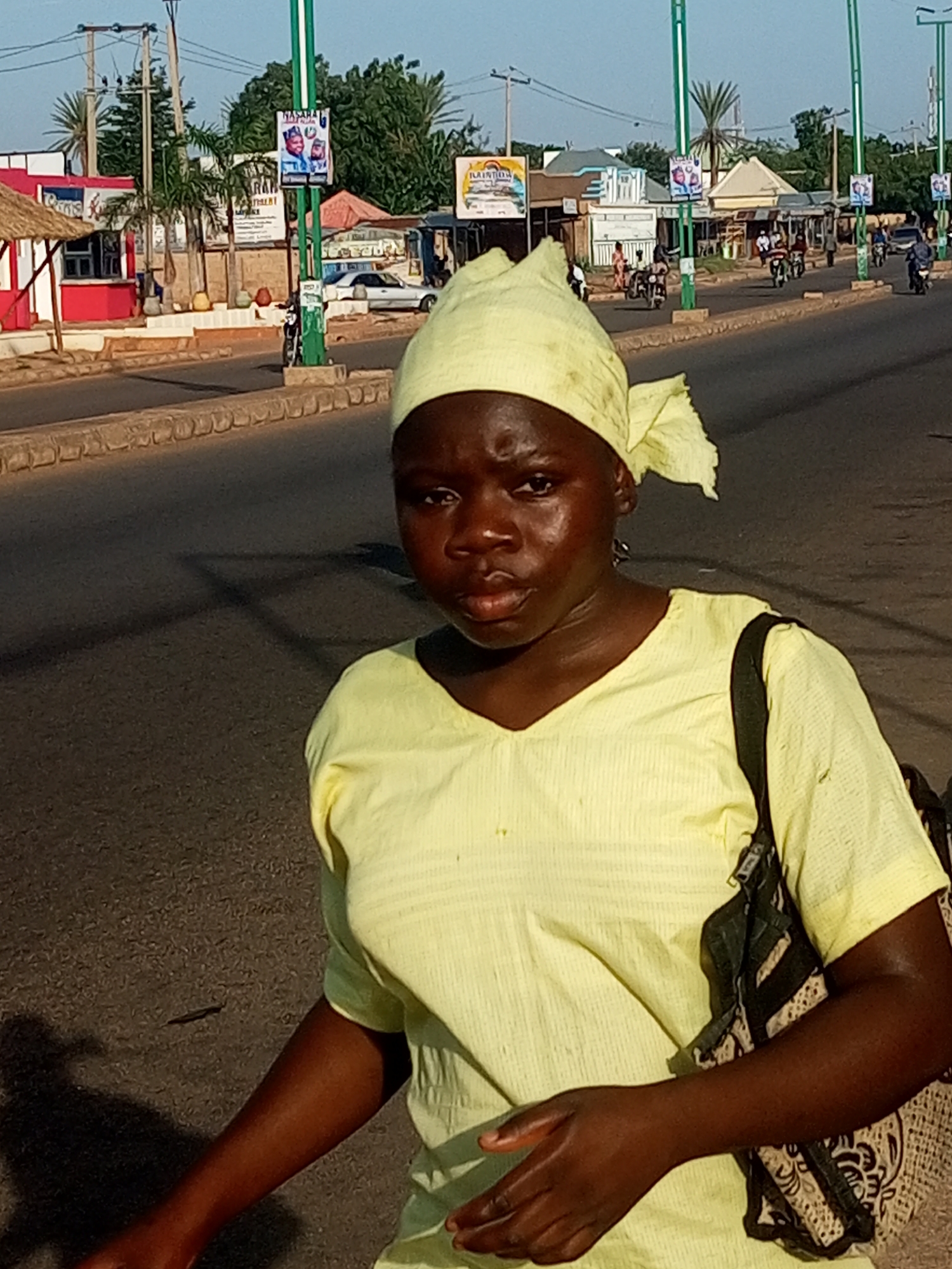 A young Black female wearing a yellow headscarf and a yellow V-neck shirt with pinstripes stands on a street in Tempure, Nigeria. She carries a patterned bag on her right shoulder. The street has a paved road, a curb, and sidewalks. In the background, there are buildings with awnings, signs, and palm trees. Several motorcycles are visible on the road and sidewalk, and a car is parked further down. The sky is clear and blue, indicating it is daytime.
