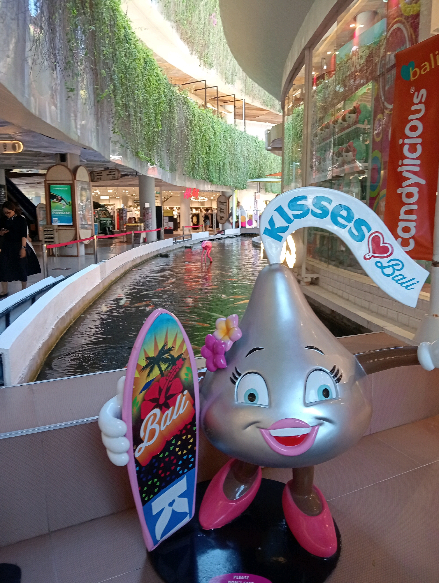 A life-sized silver Hershey's Kiss mascot character stands in the foreground of a mall, holding a surfboard that reads "Bali". The character has large blue eyes, pink lips, and is wearing pink ballet flats. Behind the mascot is an indoor koi pond, with a pink flamingo statue visible in the water. The mall features lush greenery hanging from upper levels and a Candylicious store in the background, suggesting the location is in Kuta, Indonesia. A banner for Candylicious Bali is visible on the right. Several shoppers are visible in the background.