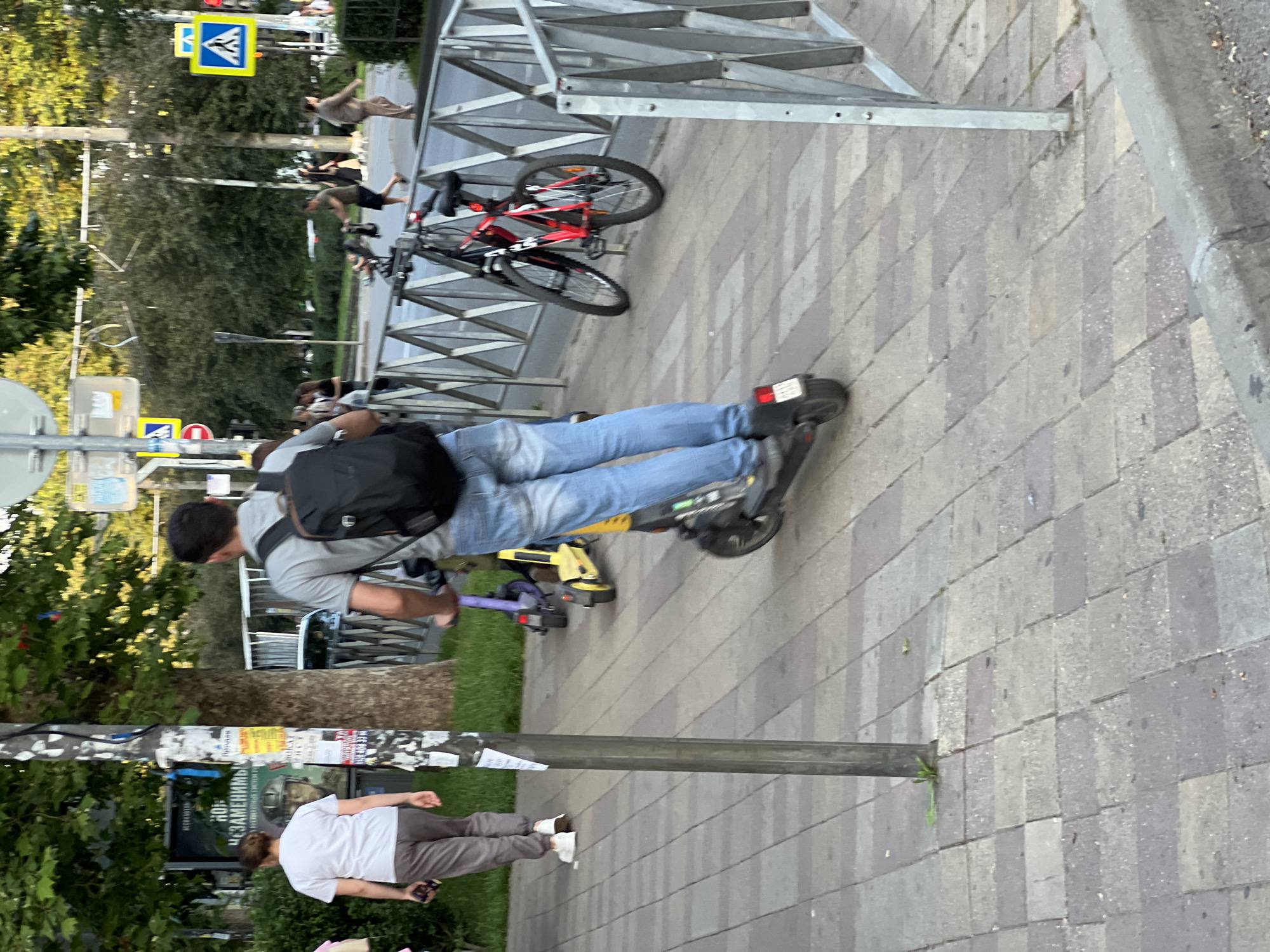The image depicts a sidewalk scene in Krasnodar, Russia. A man is riding a black electric scooter, wearing a gray t-shirt, blue jeans, and a black backpack. To his left, a woman in a white t-shirt and gray pants walks away from the camera, holding a phone. In the background, a red and black bicycle is parked near a metal fence. The sidewalk is paved with light-colored tiles, and a metal pole stands near the woman. Trees and greenery line the background, with a street and more pedestrians visible further down. The overall setting appears to be an urban environment during daylight hours.