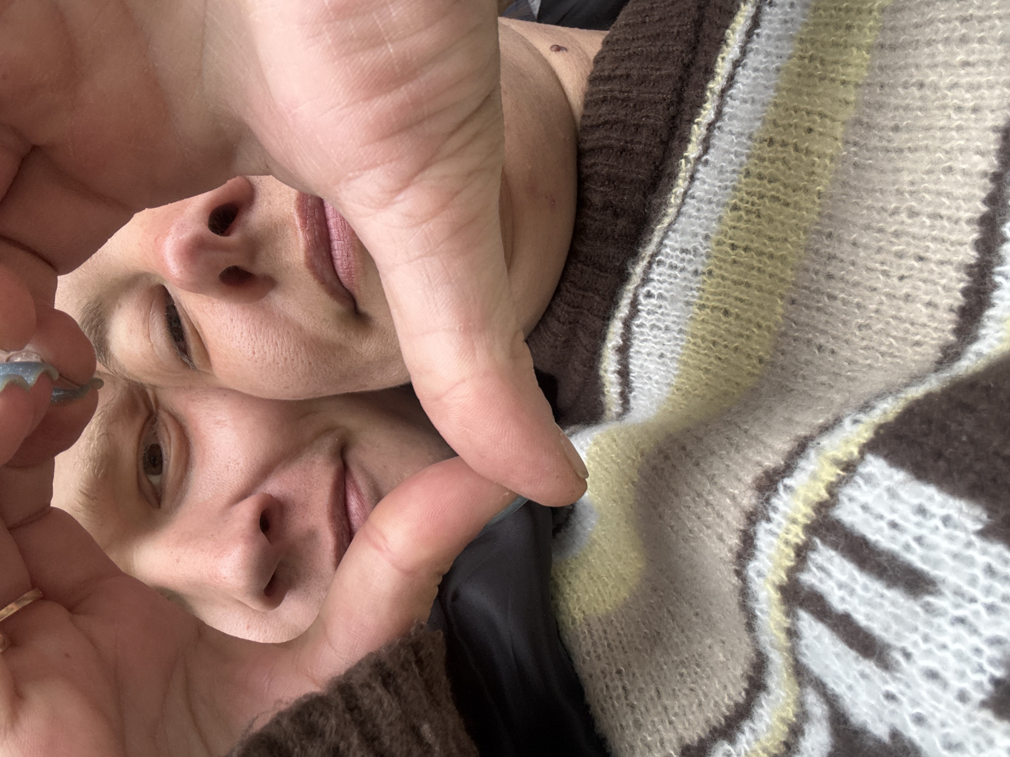 A close-up, upside-down selfie shows two people with light skin. One person is positioned at the top of the frame and the other at the bottom, with a hand partially obscuring both faces. The top individual has closed eyes and slightly parted lips, while the bottom individual has one eye open and is smiling slightly. The hand holding the camera has rings on the fingers and is framing both faces. Both individuals are wearing a multi-colored, knitted sweater. The background is mostly filled with the sweater. The location and context of the city of Dmitrov, Russia, cannot be determined from the visual data provided.