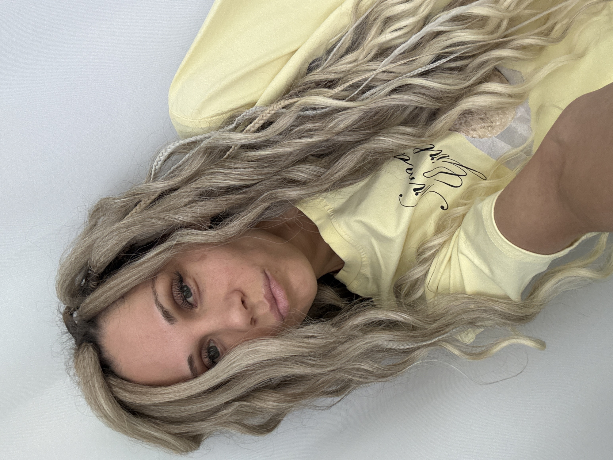 A woman with long, wavy, blonde and silver braids is lying on a white surface. She is wearing a pale yellow long-sleeved shirt with black writing on the front. Her face is turned towards the camera. The background is a plain white surface, possibly a bedsheet or similar fabric. The location is not discernible from the image.