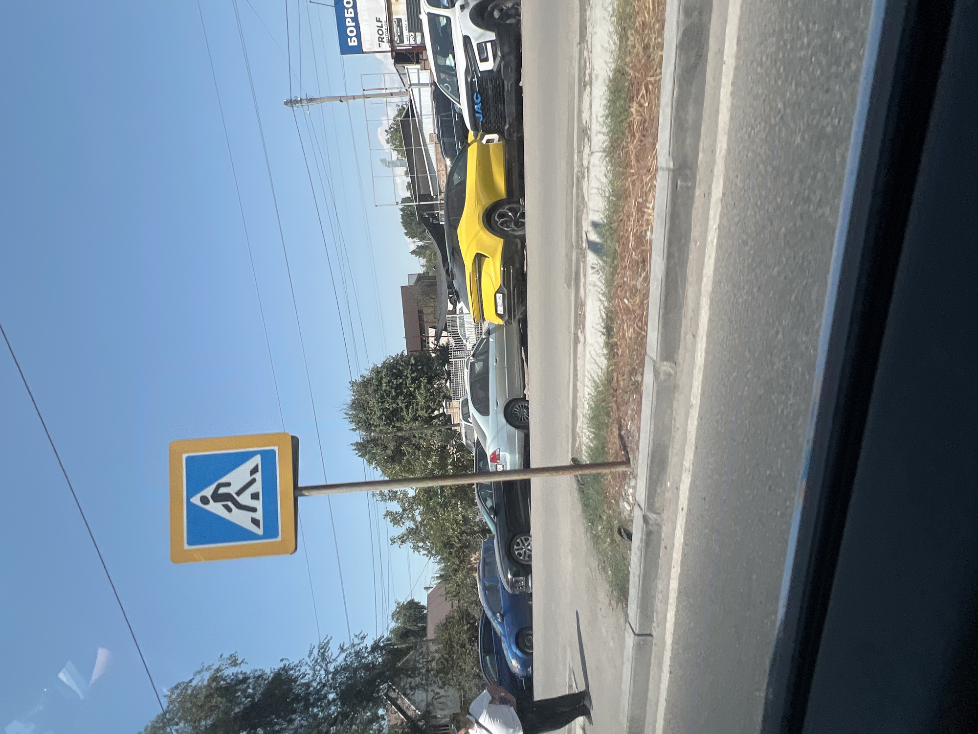 The image captures a street scene in Bishkek City, Kyrgyzstan, on a sunny day. A prominent pedestrian crossing sign, featuring a blue square with a white triangle and a black pedestrian silhouette, stands on a pole in the foreground. Beyond the sign, a line of parked cars is visible, including a bright yellow sports car, a white pickup truck with a "JAC" logo on its grille, and several other vehicles in various colors. A person in a white shirt and dark pants stands on the sidewalk on the left side of the image, facing away from the camera. The background shows buildings and trees under a clear blue sky. The perspective suggests the photo was taken from a moving vehicle, looking towards the right.