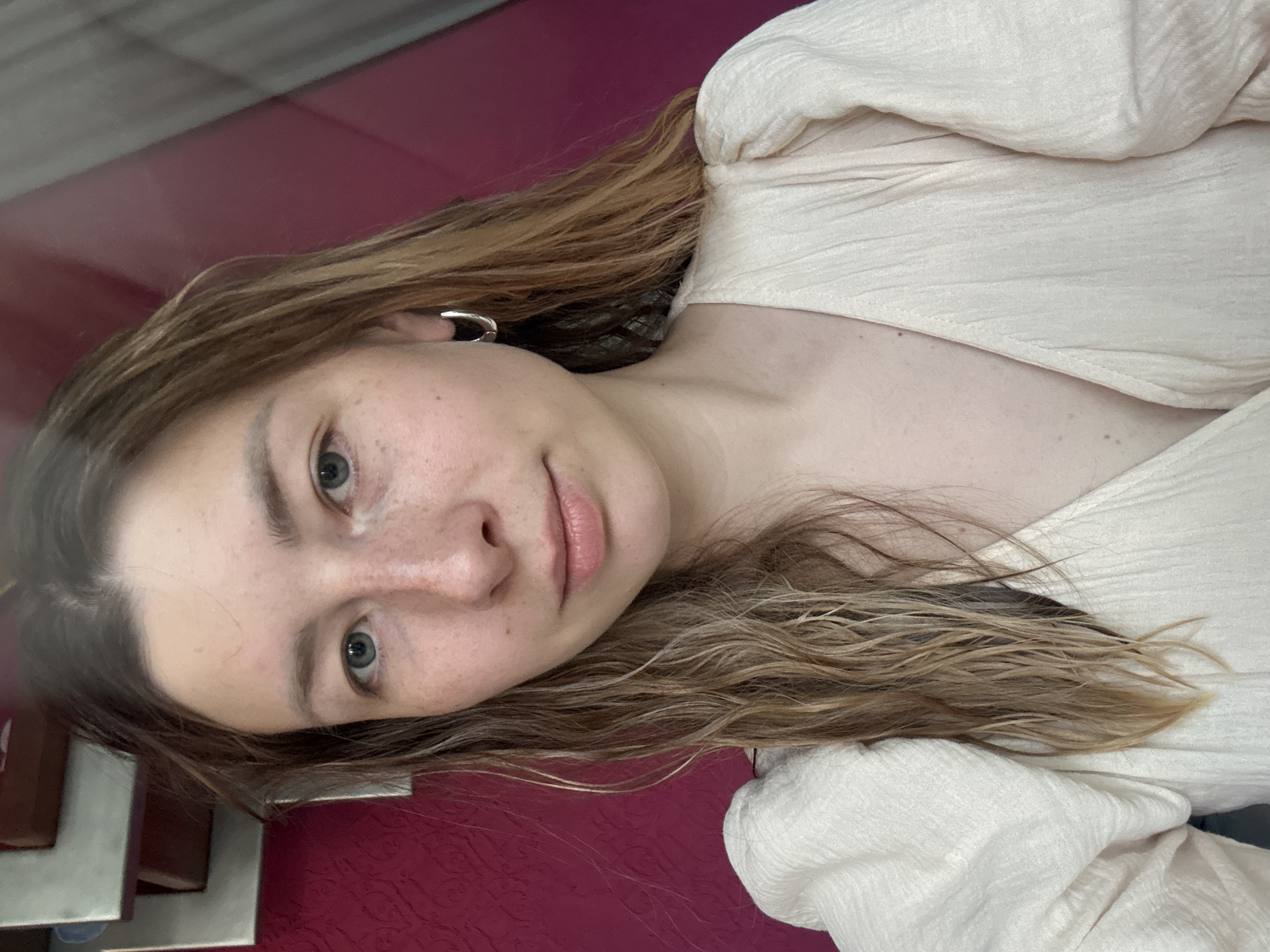 A young woman with light skin and brown hair is depicted in a close-up portrait. She has freckles on her face and is wearing a light-colored, textured shirt. The background consists of a maroon surface and an out-of-focus, undefined object. The location of Чёрное, Russia, is not discernible from the image.