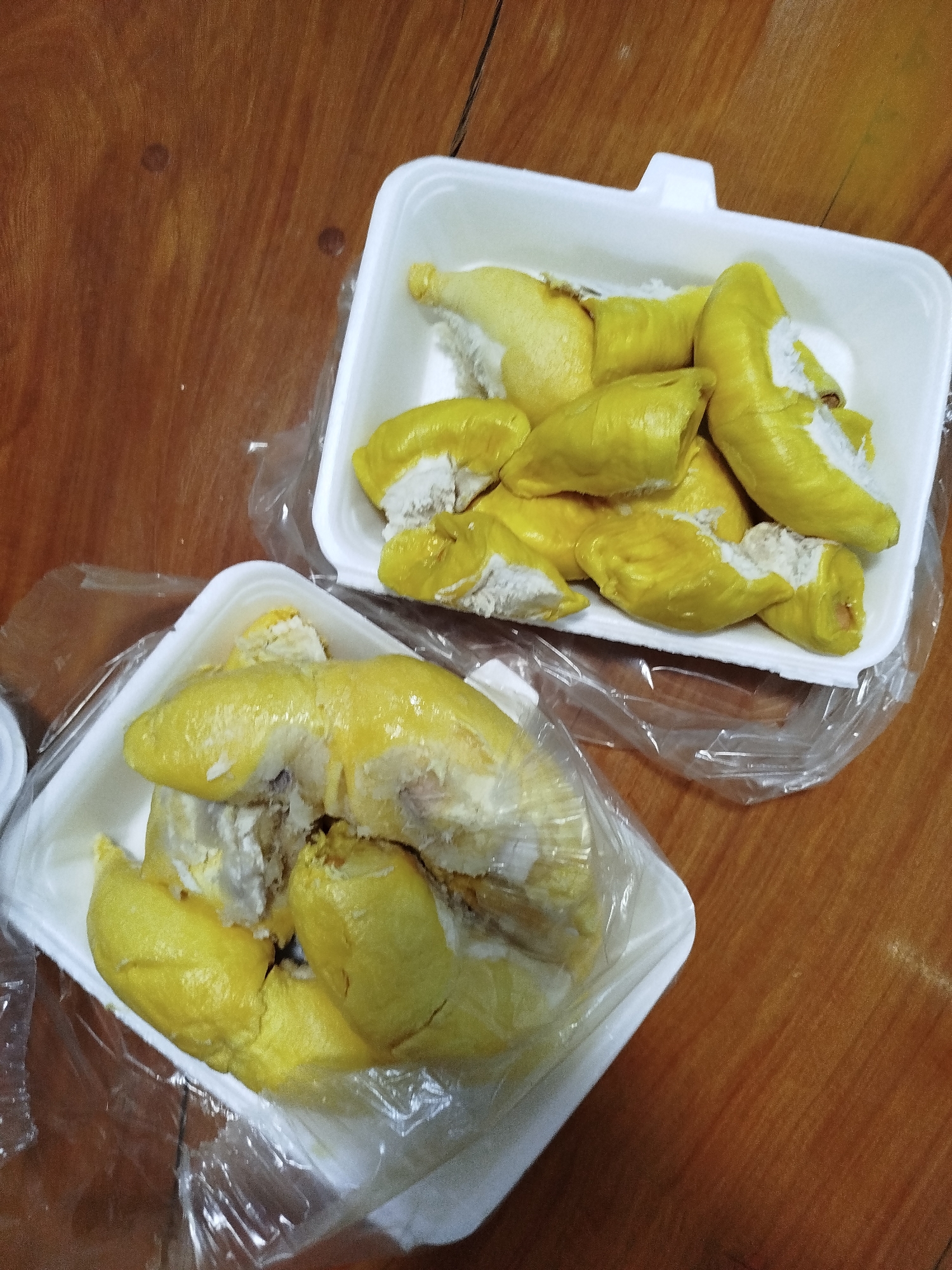 Two white styrofoam containers are placed on a wooden table. Each container contains segments of durian fruit. The durian segments are yellow and fleshy, some with white pulp visible. The containers are partially covered with clear plastic wrap. The table is made of brown wood with a smooth, possibly varnished, surface. The scene appears indoors. There is no evidence to confirm it is in Da Lat, Vietnam.