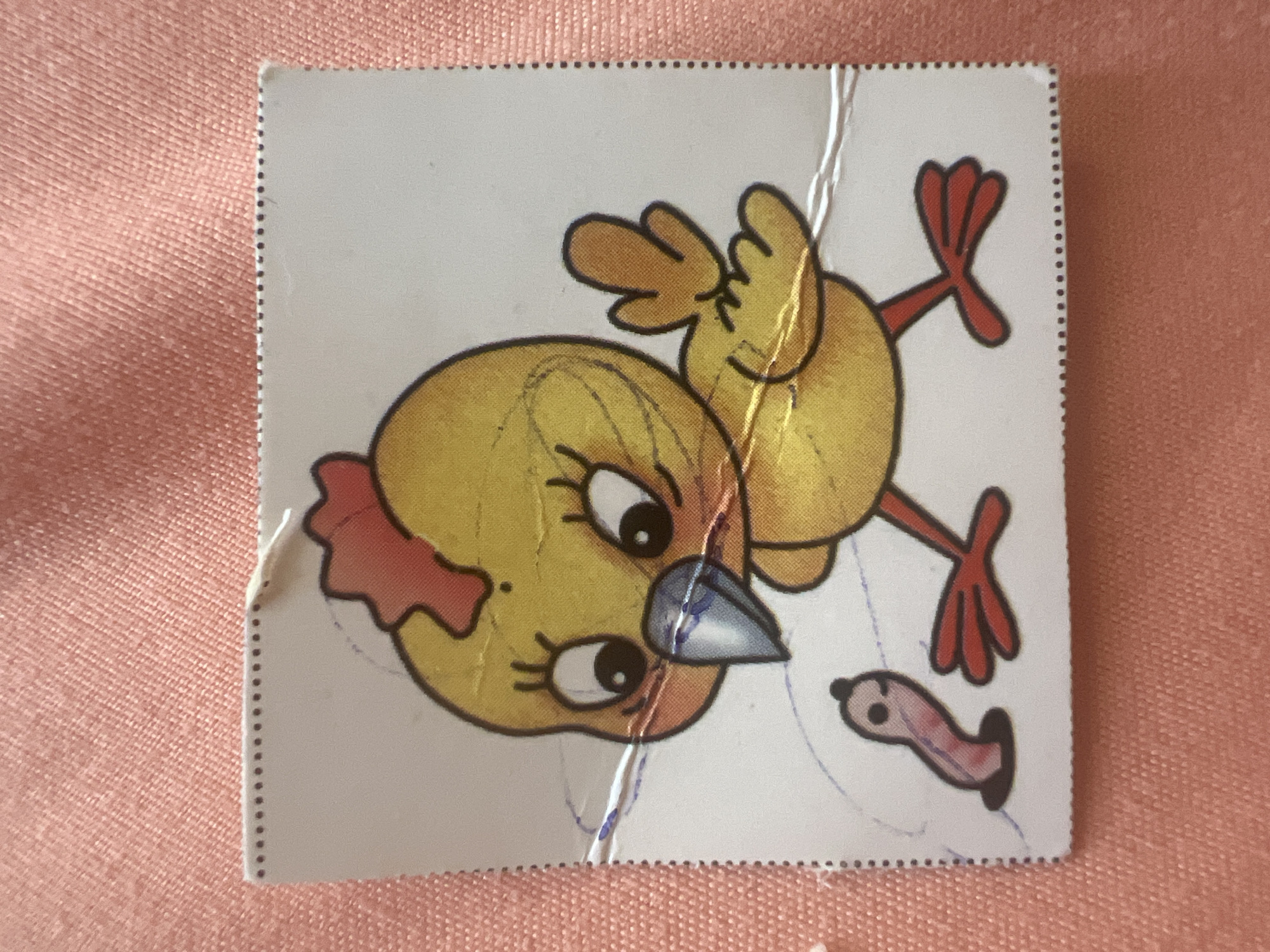 The image is a close-up, high-angle shot of a small sticker featuring a cartoon chick and a worm on a pink, textured surface. The sticker is square with a perforated edge along the top and right sides. The chick is yellow with a red crest, large black eyes with eyelashes, and a blue and gray beak. It has orange feet. A small, pink worm with a black head is emerging from a dark brown hole on the left side of the sticker. The sticker appears to be slightly damaged, with some creases and what looks like faint blue scribbles on it. The pink surface underneath is consistent with fabric. There is no information in the image to suggest any connection to the city of Pskov, Russia.