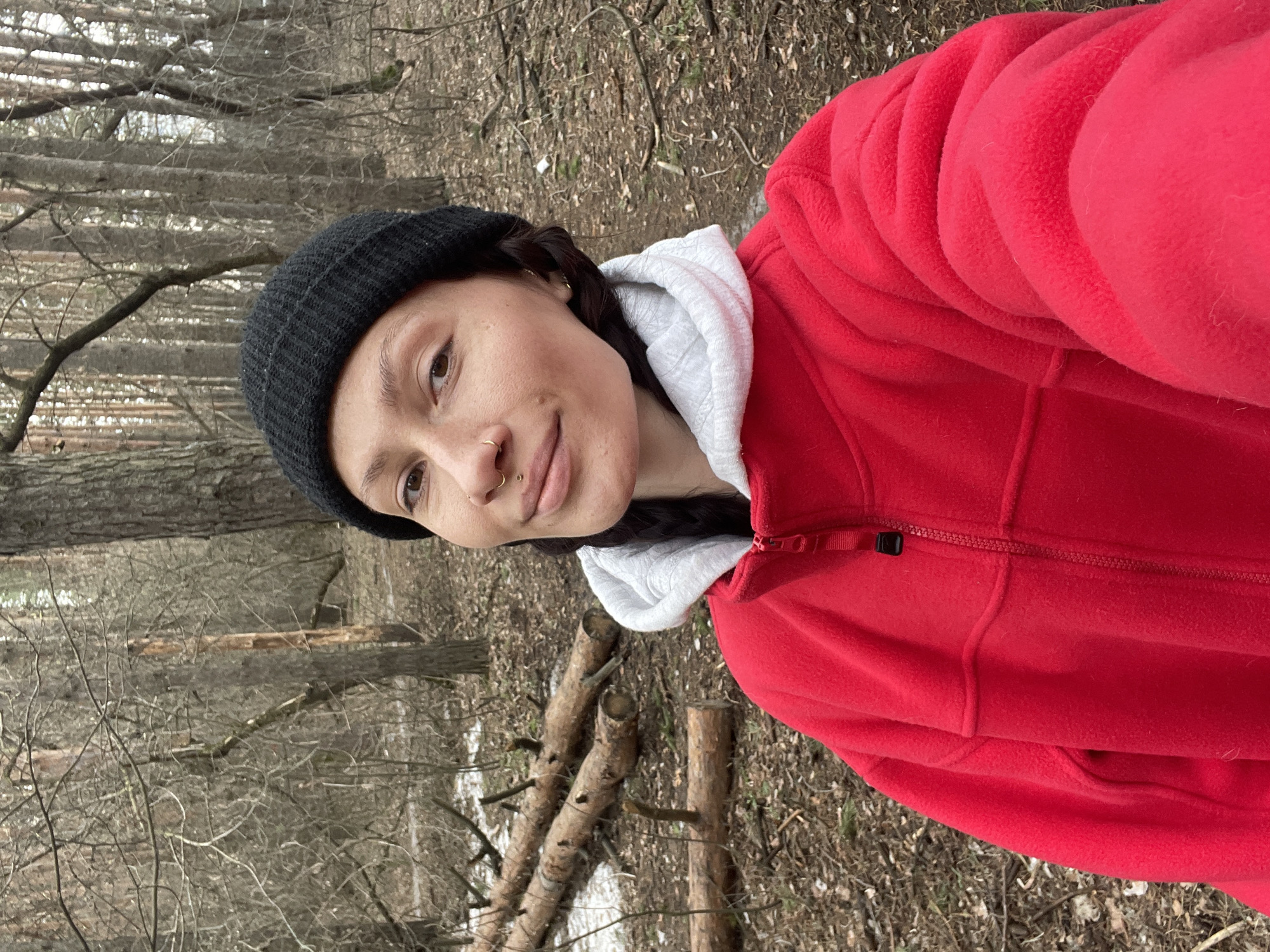 A woman is standing in a forest. She is wearing a black beanie, a white hoodie, and a red fleece jacket. The forest has bare trees and fallen logs. The ground is covered with leaves and some patches of snow. The location appears to be a wooded area, possibly on the outskirts of a city, but there is no definitive information in the image to confirm it is Izhevsk, Russia.