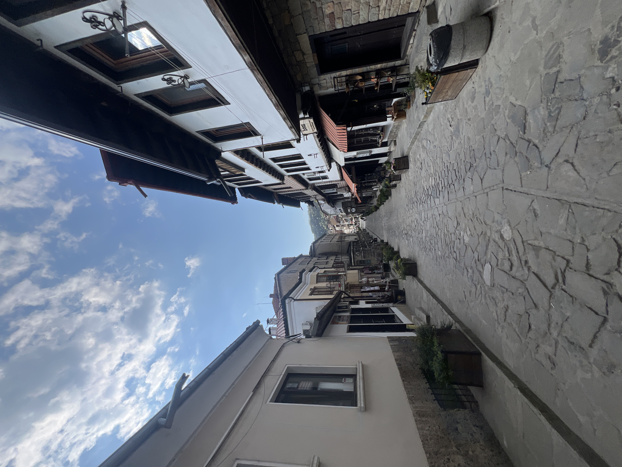The image captures a sunny, daytime view down a narrow, historic street or alley, characteristic of Veliko Tarnovo, Bulgaria. The stone-paved street, composed of irregular, light-colored slabs, recedes into the distance, flanked by multi-story buildings on both sides.
On the left, the buildings feature white facades with dark wooden beams and window frames, some with overhanging upper stories. Ornate black metal light fixtures or decorations hang from the upper levels. Reflections of the bright blue, cloudy sky are visible in some windows. A small sign on one of these buildings clearly reads "Bar". The buildings on the right are constructed from a mix of stone and lighter-colored stucco, also featuring traditional architecture with dark-framed windows and doors, some with wooden awnings. Planter boxes with greenery are placed along the street and mounted on the buildings.
The sky is bright blue with scattered white, puffy clouds, indicating clear weather. The street appears empty of people, contributing to a peaceful and timeless atmosphere. The overall scene highlights the quaint and historical charm of the location, with traditional architecture and a well-preserved streetscape.
